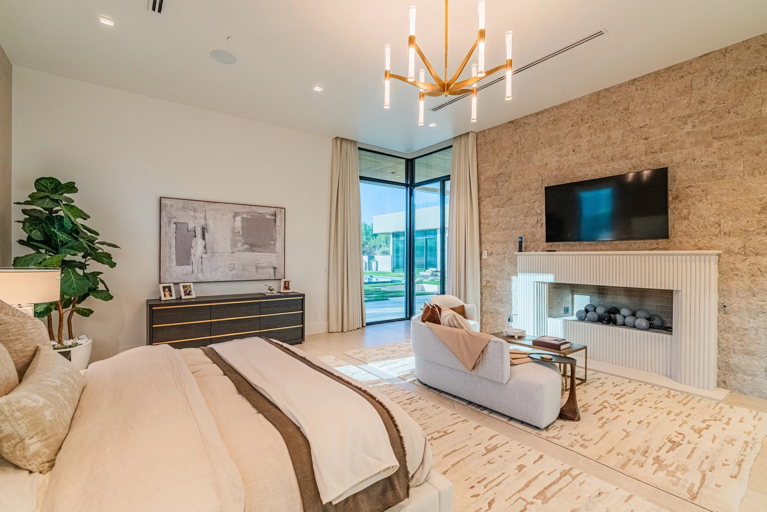 A modern living room with a beige sofa, a white armchair, a black sideboard, a large wall-mounted TV, a fireplace with decorative stones, a potted plant, large glass windows with curtains, and a contemporary chandelier.