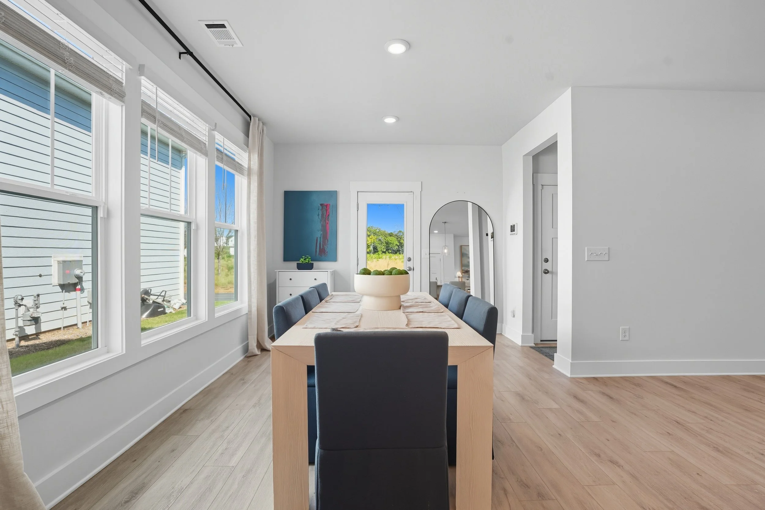 Modern dining room with large windows, a wooden table, and eight blue chairs, white walls, light wood flooring, and a colorful abstract painting on the wall.