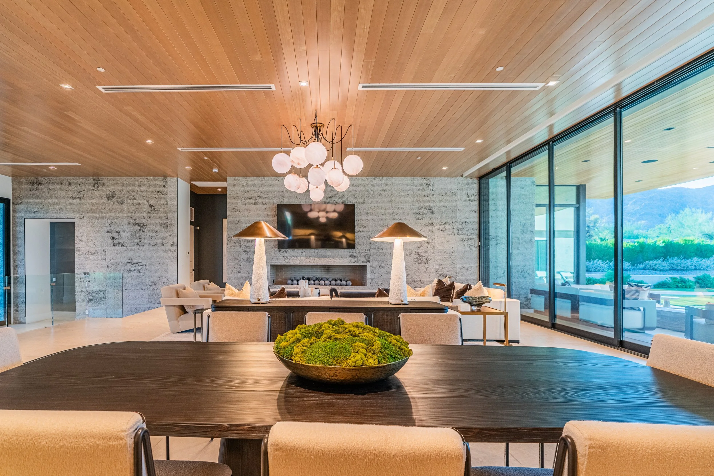 Elegant modern living room with a wood-paneled ceiling, a chandelier with multiple round glass globes, a textured brick fireplace, large windows overlooking a scenic outdoor landscape, and a dining table with a centerpiece.