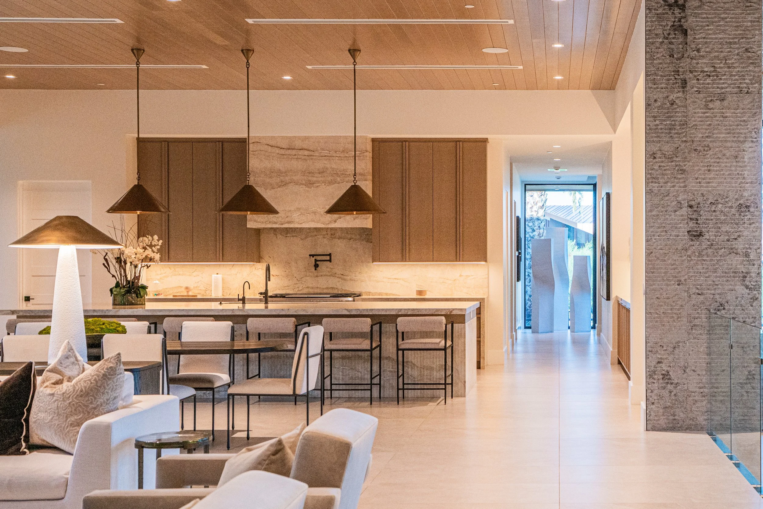 Modern kitchen with wooden ceiling, pendant lights, marble backsplash, beige cabinetry, and a view into a hallway with large windows and outdoor scenery.