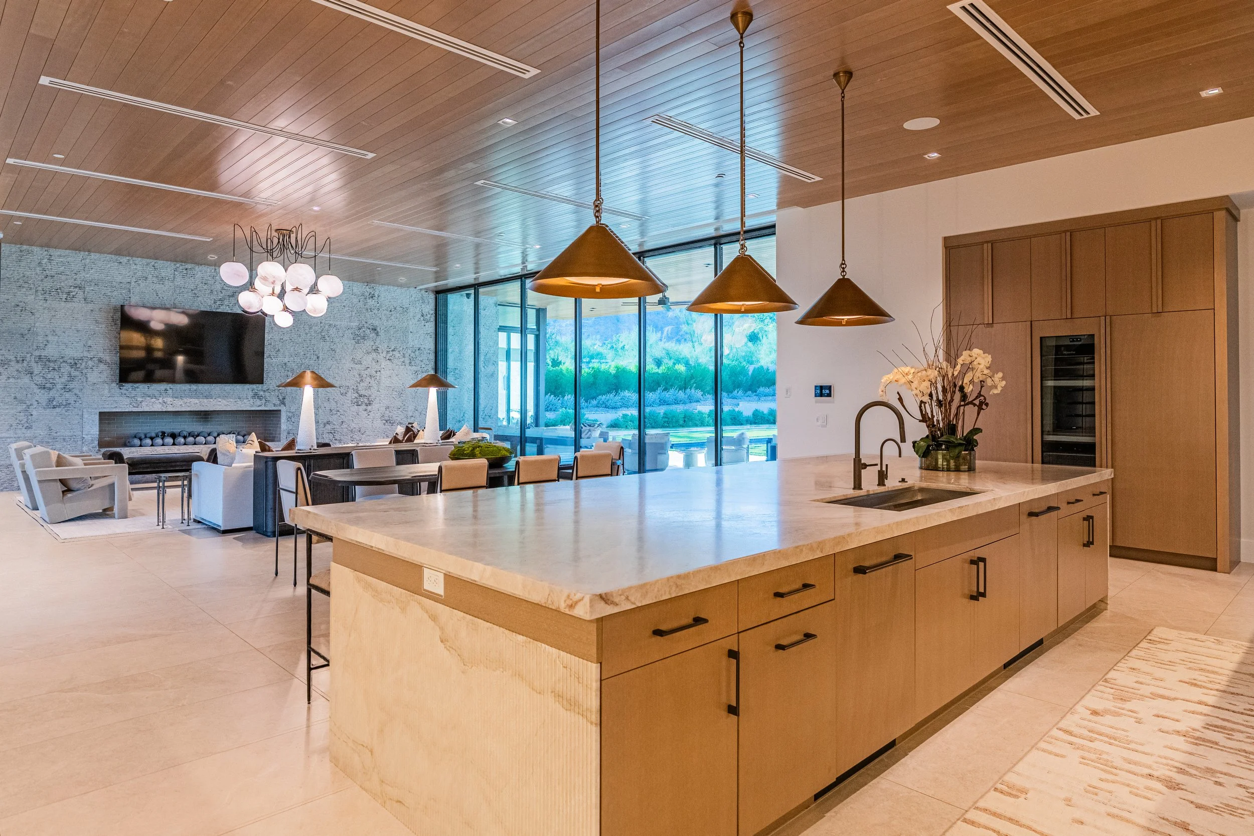 Modern kitchen with large island, pendant lights, wood ceiling, and large windows with scenic view.