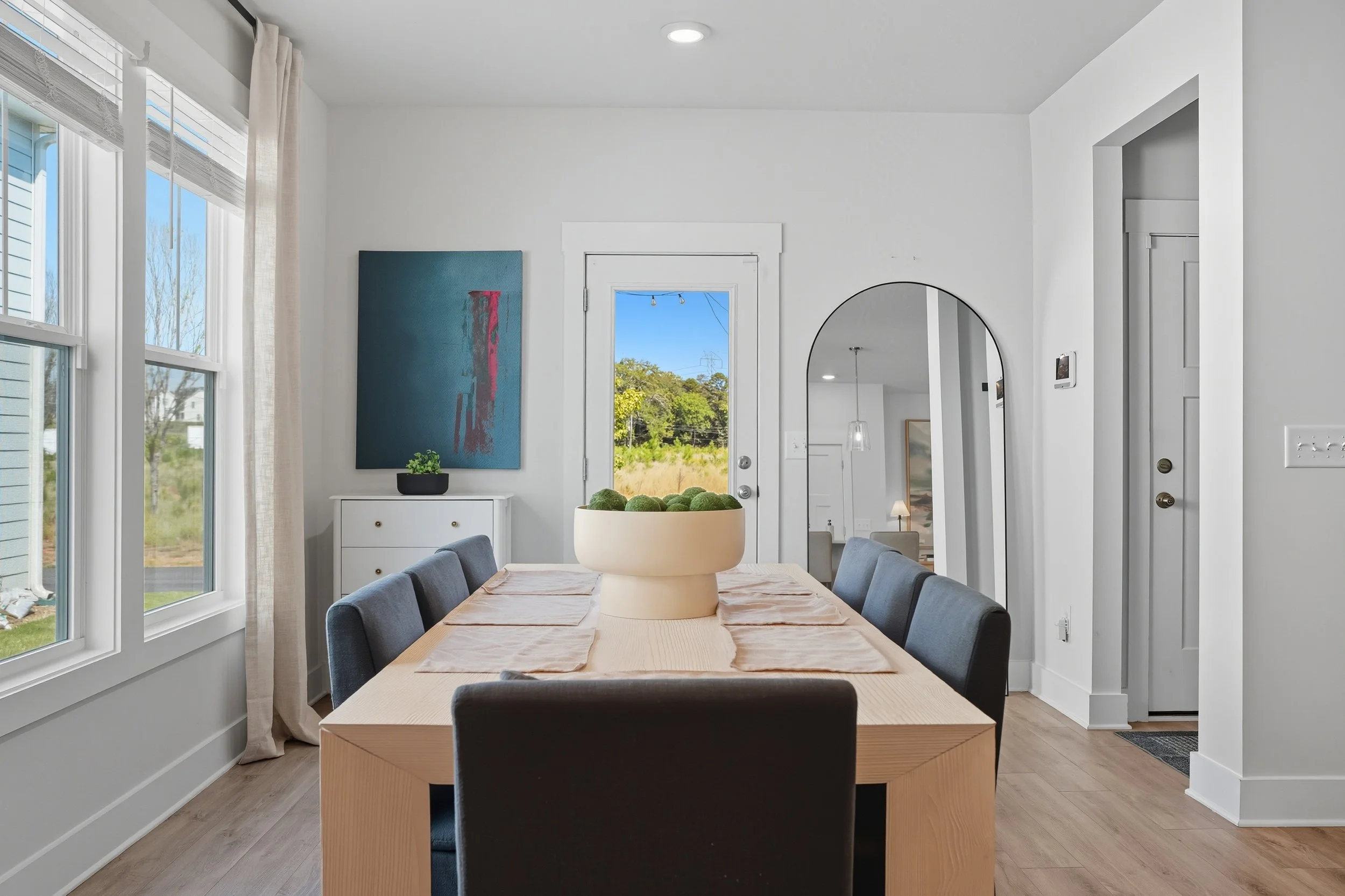 Dining room with a light wood table, six dark grey chairs, a large beige planter with green moss as a centerpiece, bright windows with white trim, and patio door overlooking a green landscape.