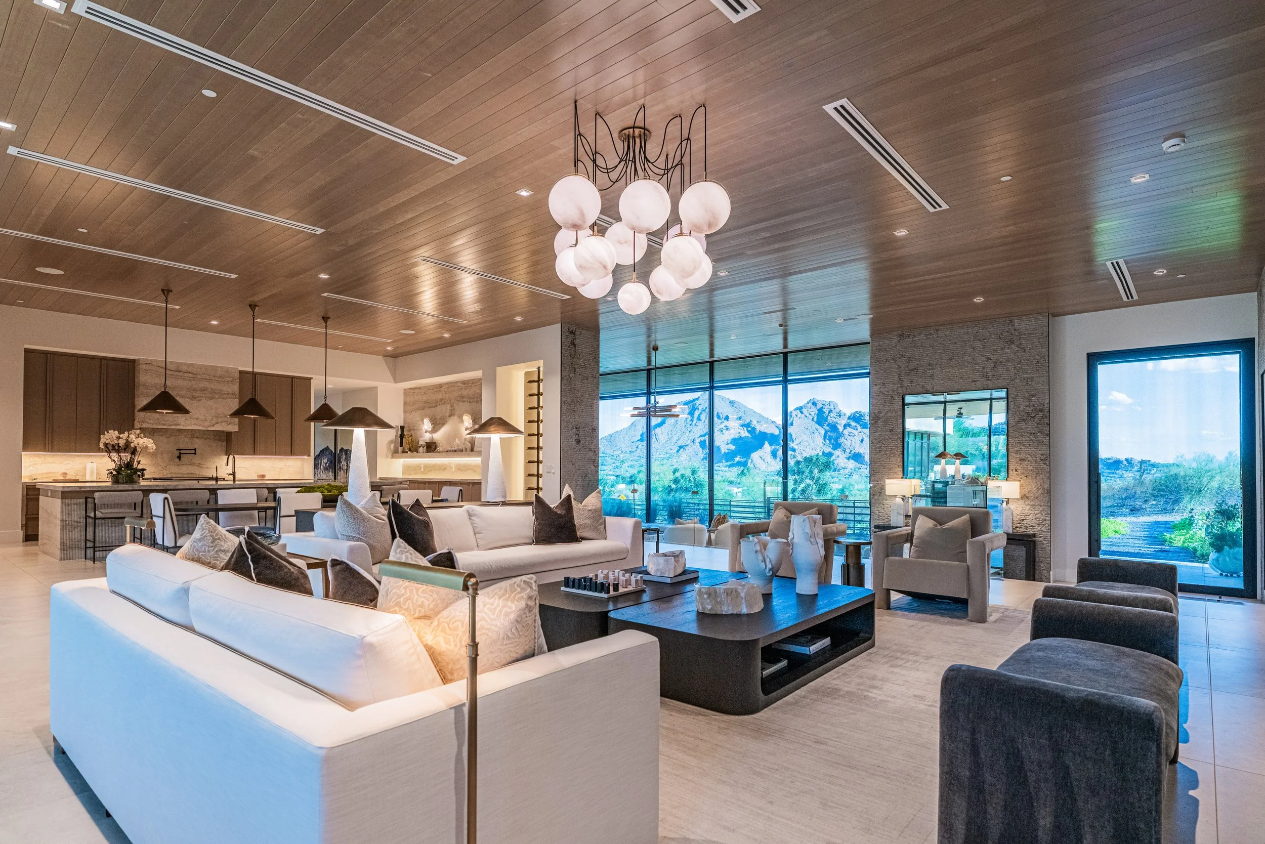 Modern living room with white sofas, brown and gray pillows, contemporary furniture, a large window with mountain view, wooden ceiling, and chandelier with white globe lights.