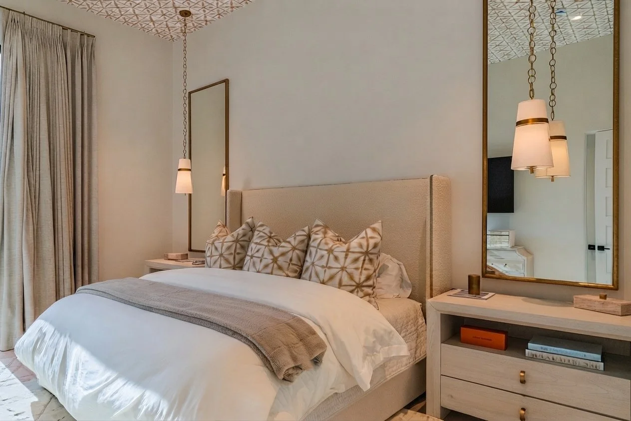 A bedroom with a large bed, beige upholstered headboard, multiple patterned pillows, and a white duvet. On each side of the bed, there are hanging pendant lights with white shades. There are large mirrors hanging on the wall behind each bedside table