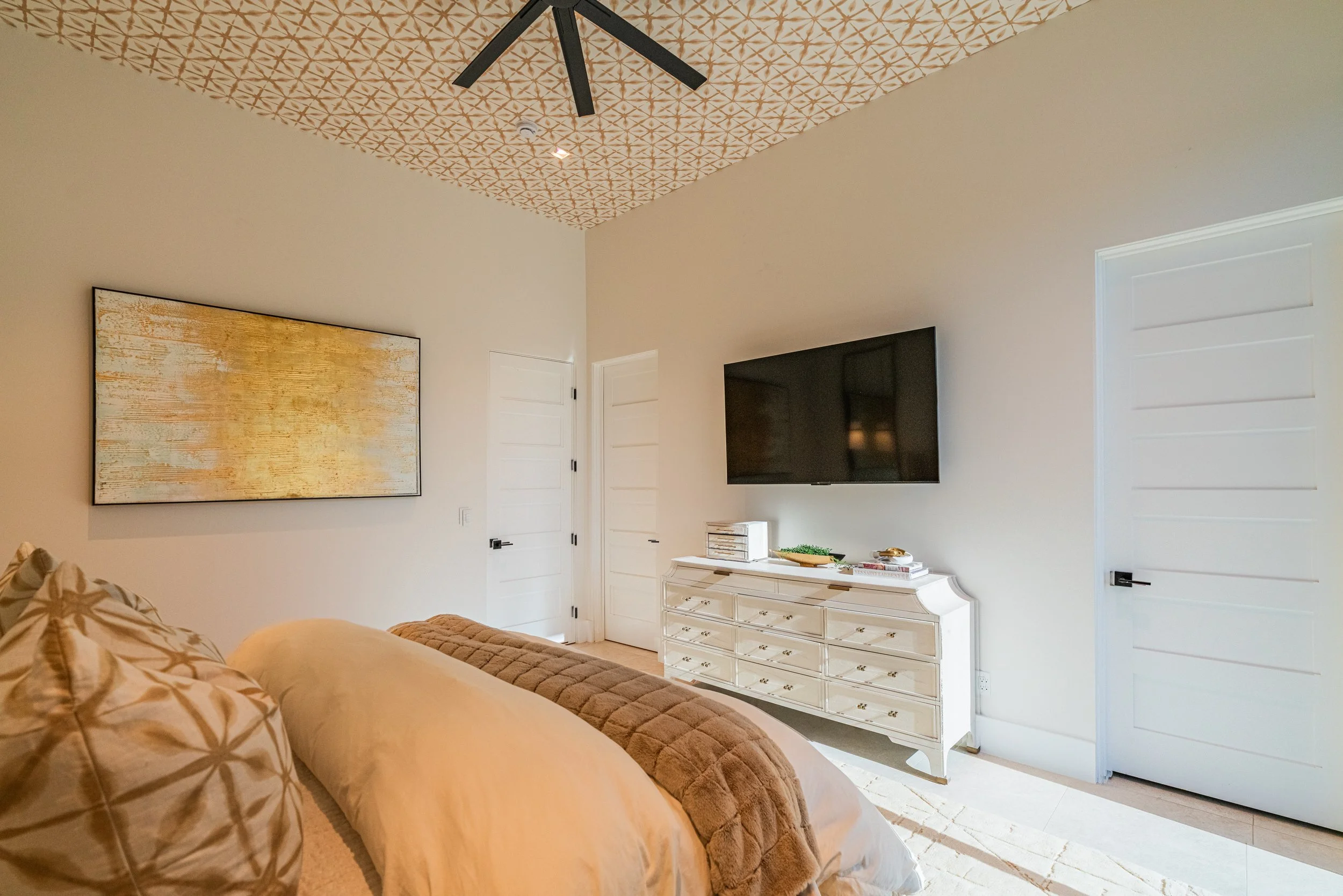 A living room with beige walls and a patterned ceiling. A large flat-screen TV is mounted on the wall above a white dresser. There is a painting with a gold and white textured design hanging on the wall. Part of a beige sofa with patterned pillows an