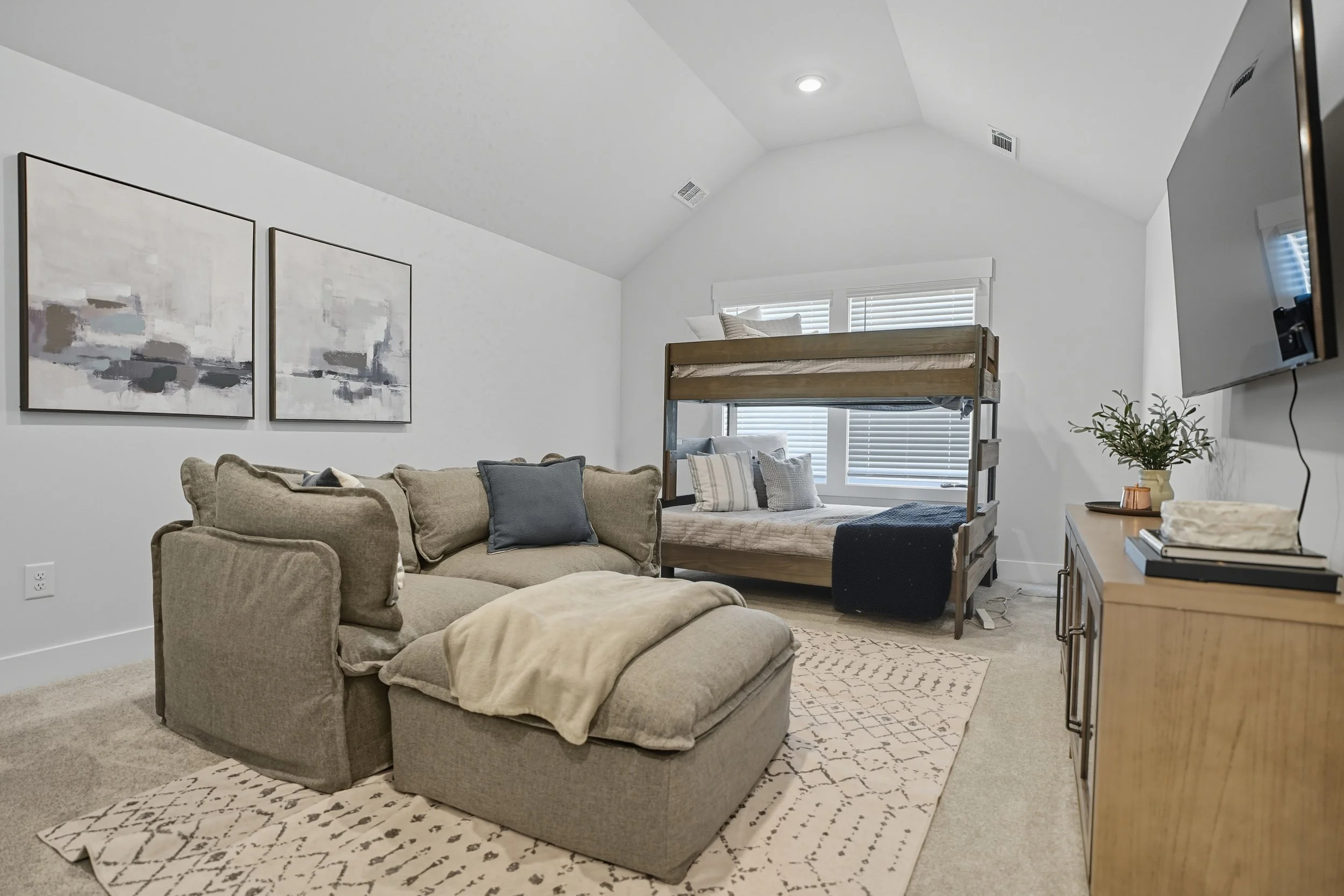Living room with a beige sofa, coffee table, patterned rug, and artwork on the wall. In the background, there is a bunk bed with pillows and blanket, and a window with blinds. A TV is mounted on the wall to the right, and a small plant is on a side t