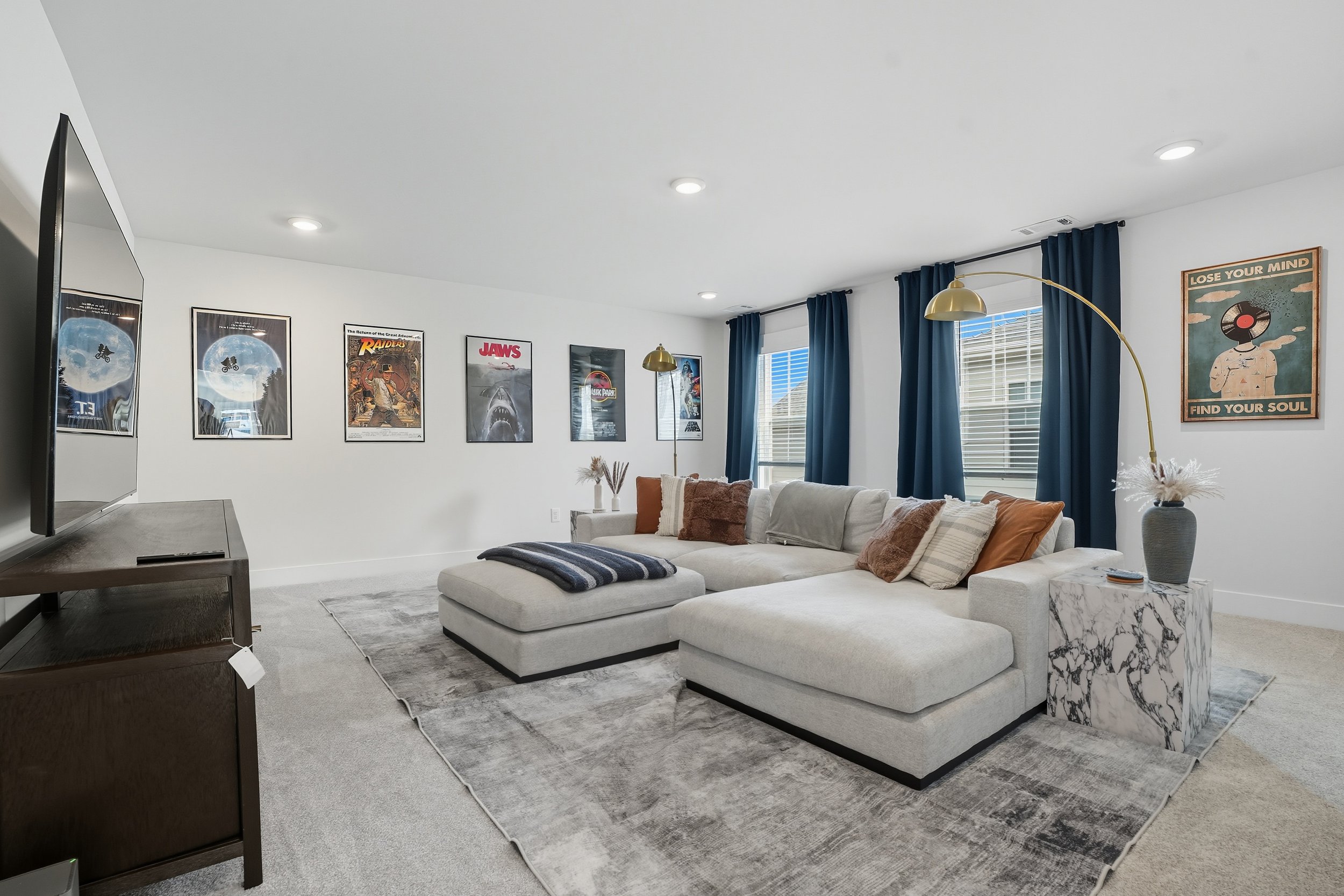 A living room with a white sectional sofa decorated with brown, beige, and striped pillows, a marble side table, and a large TV on a wooden stand. The wall is adorned with framed posters of movies and a vintage-style poster with the text 'Lose Your M