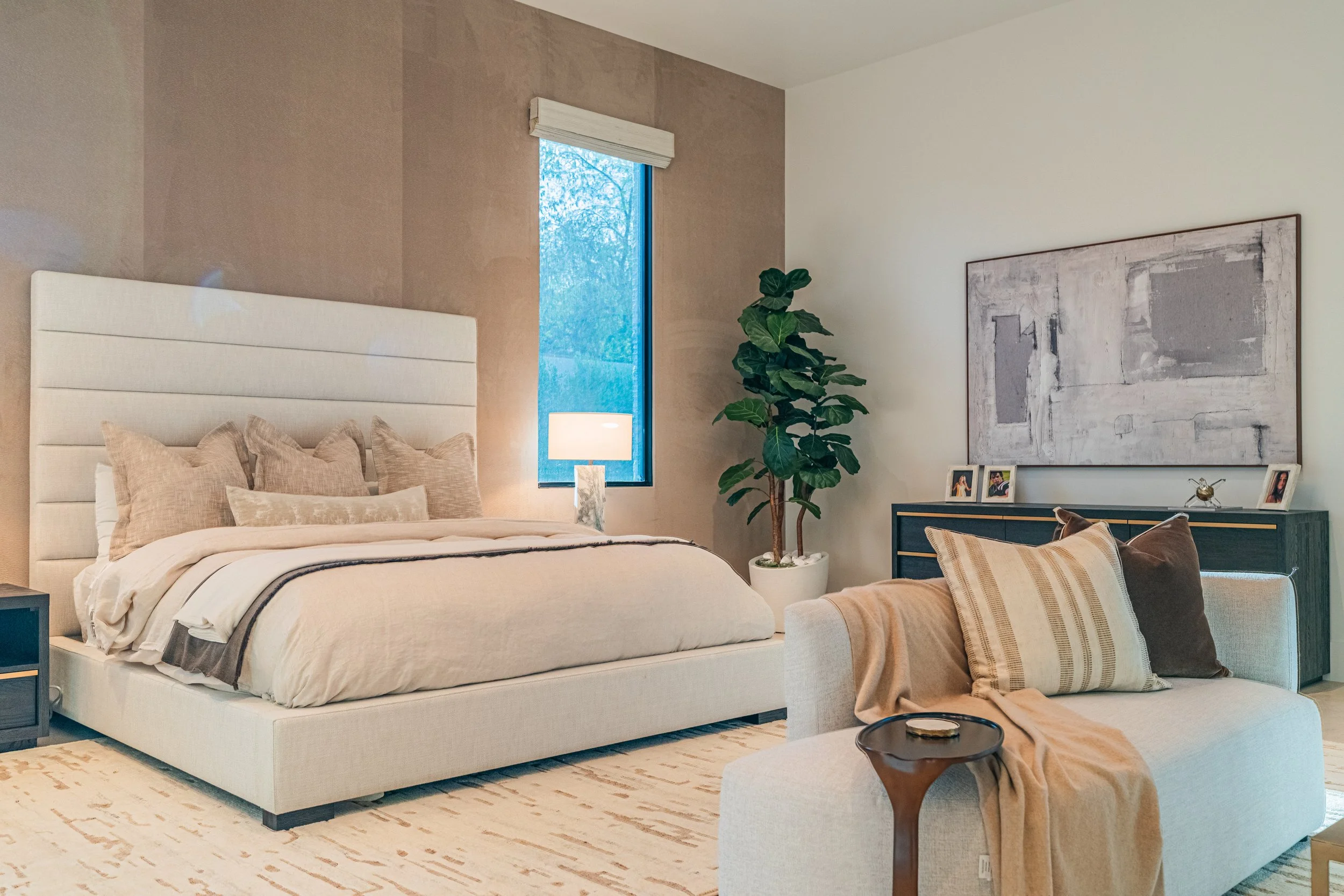A contemporary bedroom with a large bed featuring beige pillows and a white headboard, a nightstand with a lamp, a tall green plant, a large abstract painting on the wall, and a cozy seating area with cushioned chairs and throw pillows.