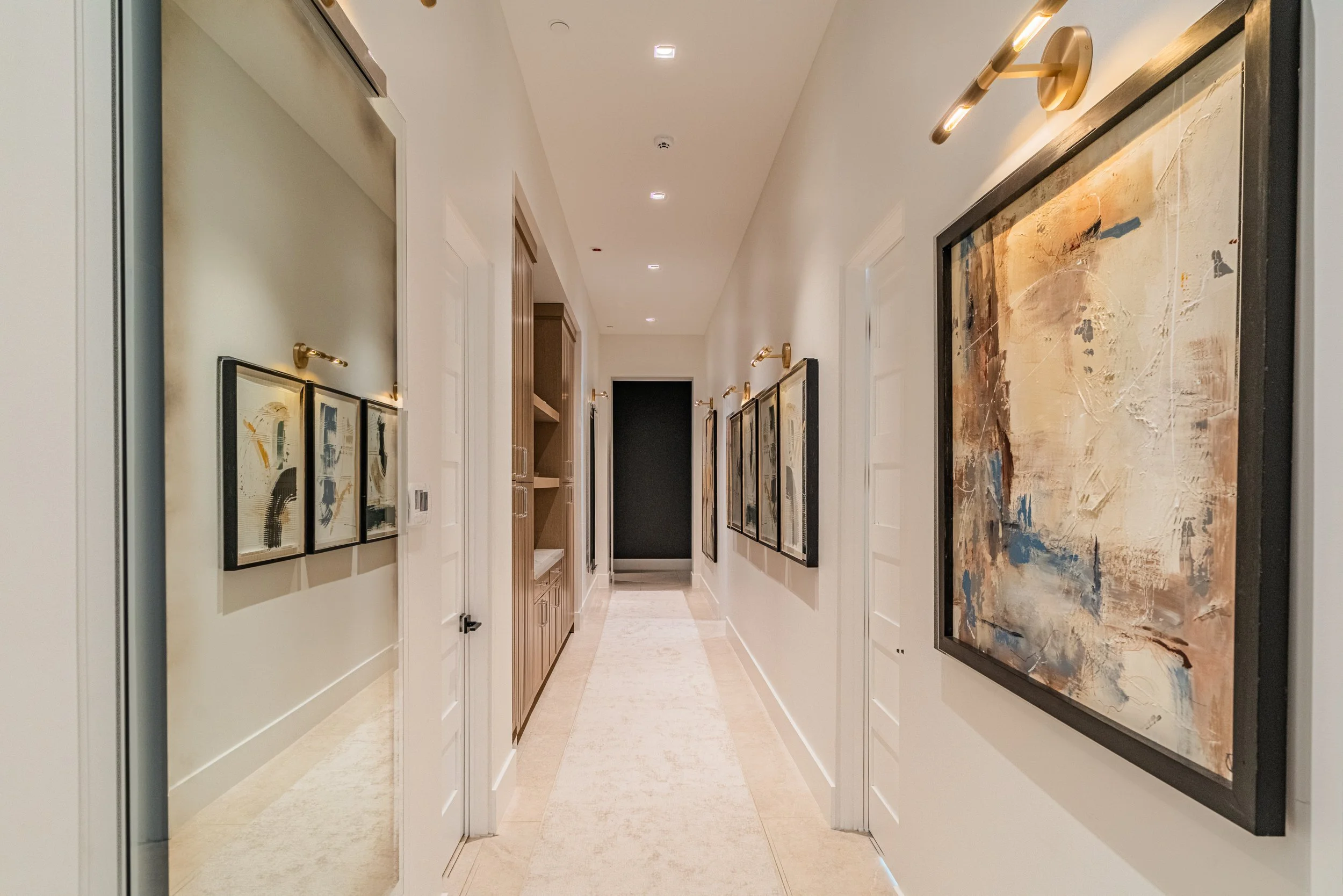 A hallway decorated with abstract art paintings on the walls, leading to a dark doorway at the end, with ceiling lights illuminating the space.