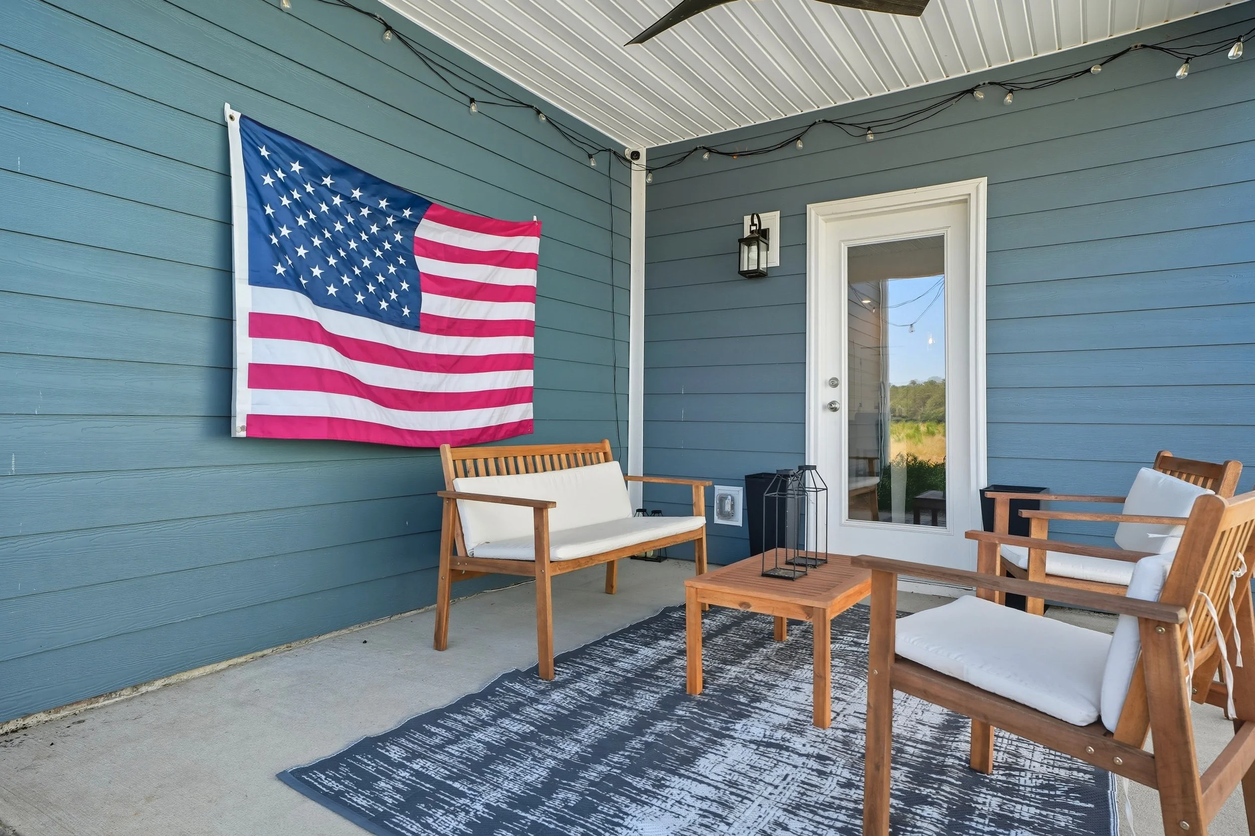 Outdoor patio with wooden chairs, a rug, and a blue wall with a large American flag hanging. String lights are draped along the ceiling, and there is a white door with a window.