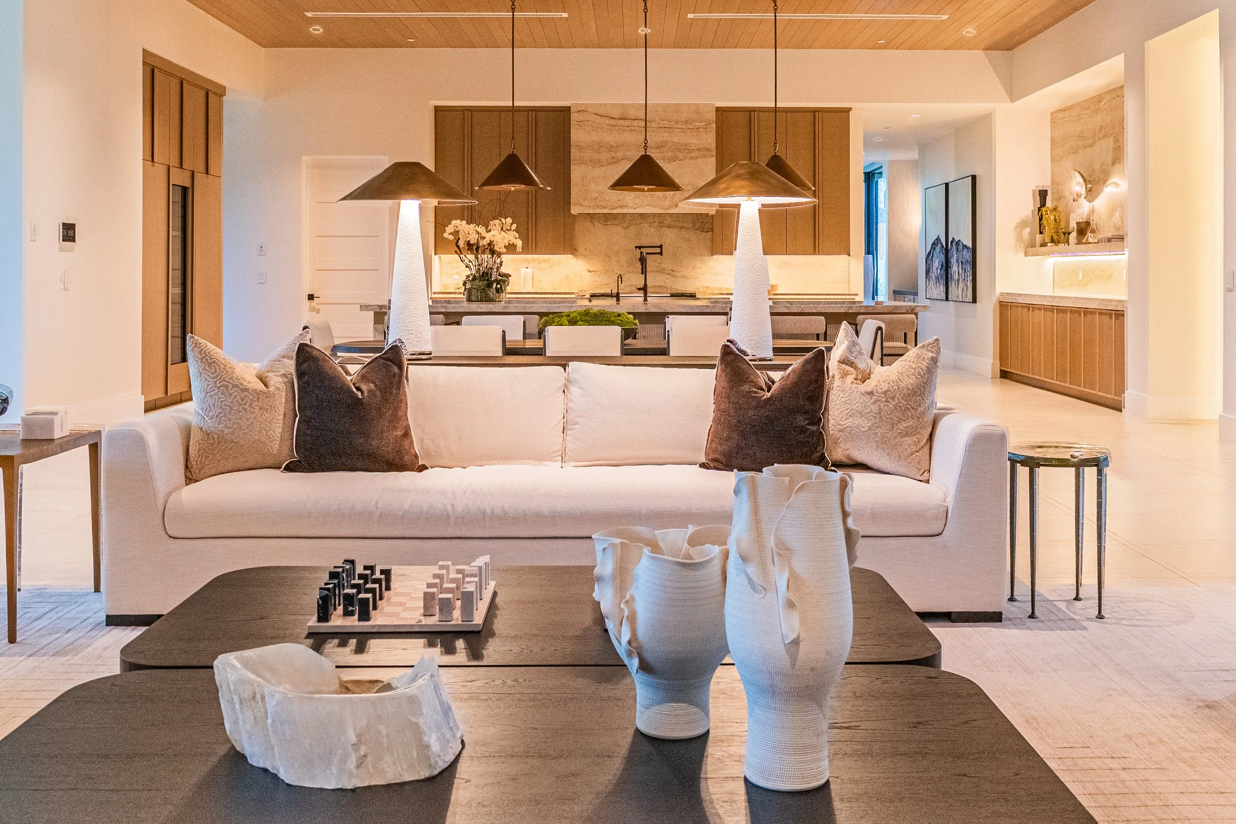 Living room with a wooden ceiling, beige walls, and contemporary furniture. Features a white sofa with brown pillows, a dark wooden coffee table with a chessboard, and decorative white ceramic vases on a table with tissue paper. In the background, th
