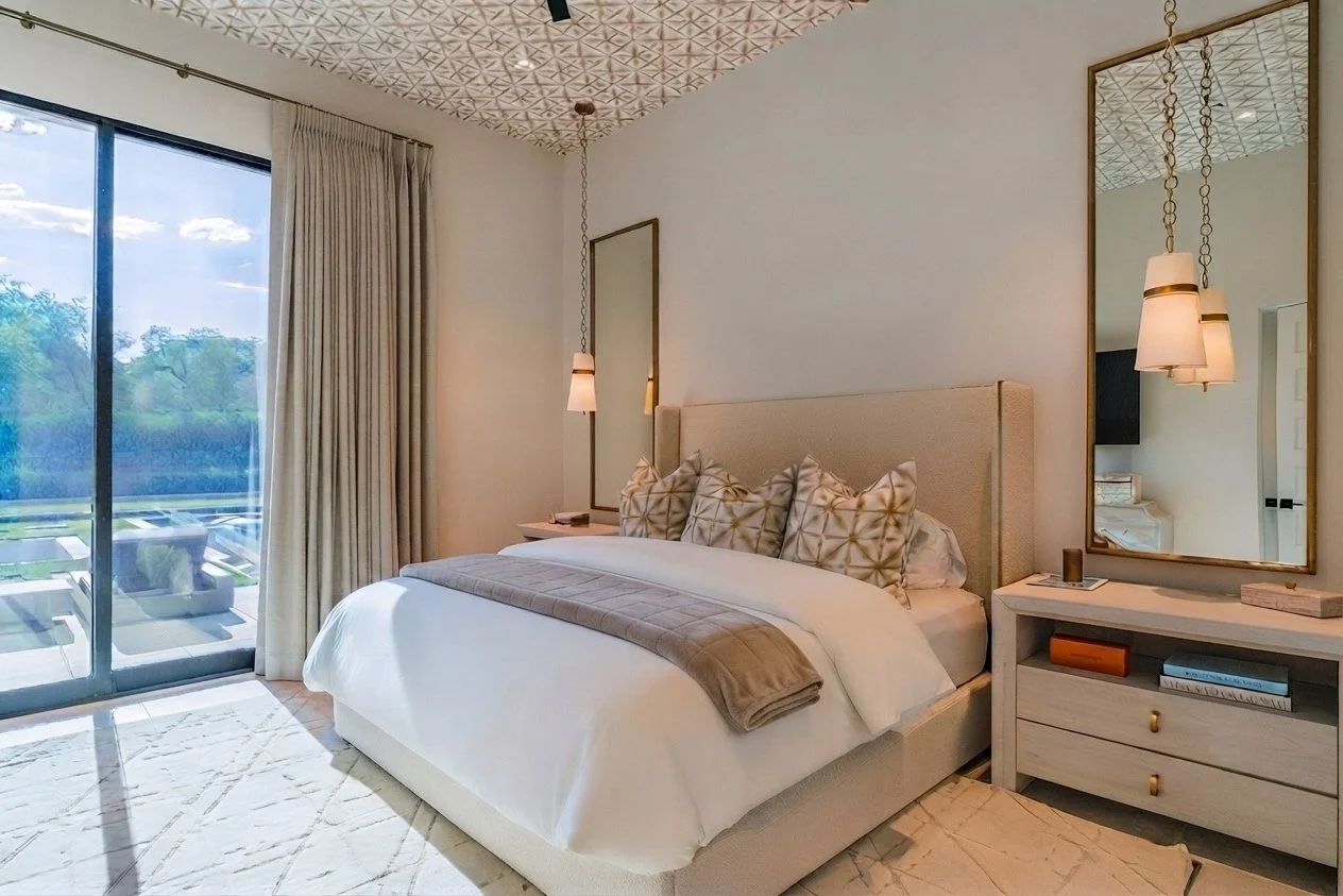 A modern bedroom with a sliding glass door leading to an outdoor balcony with chairs, a large bed with multiple decorative pillows, beige curtain, and a white dresser with a large mirror, hanging pendant lights, and a view of greenery outside.