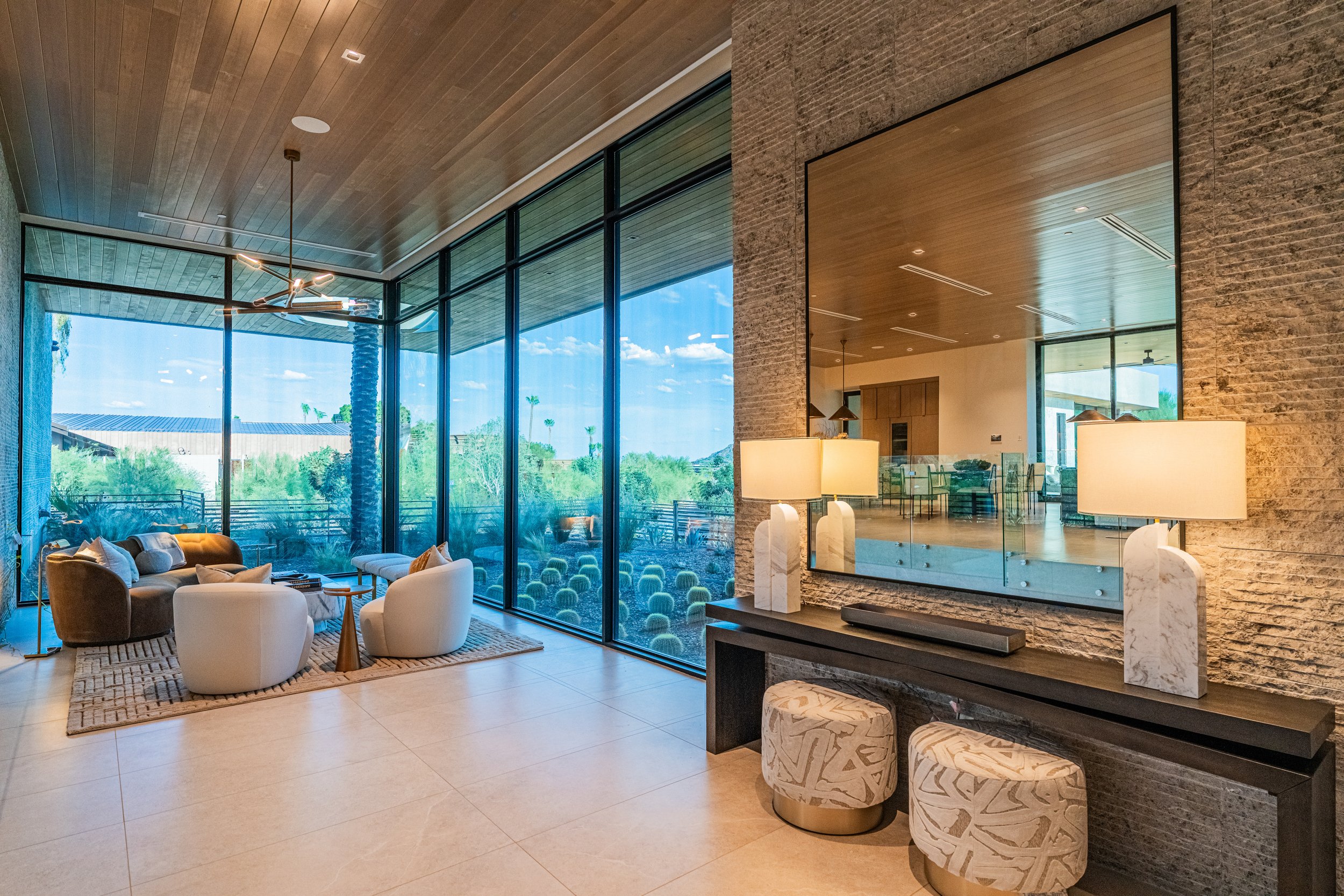 Modern living room with large floor-to-ceiling windows showing a sunny outdoor landscape with trees and a building, minimalist beige and white furniture, a textured brick wall, a large mirror, and warm lighting.