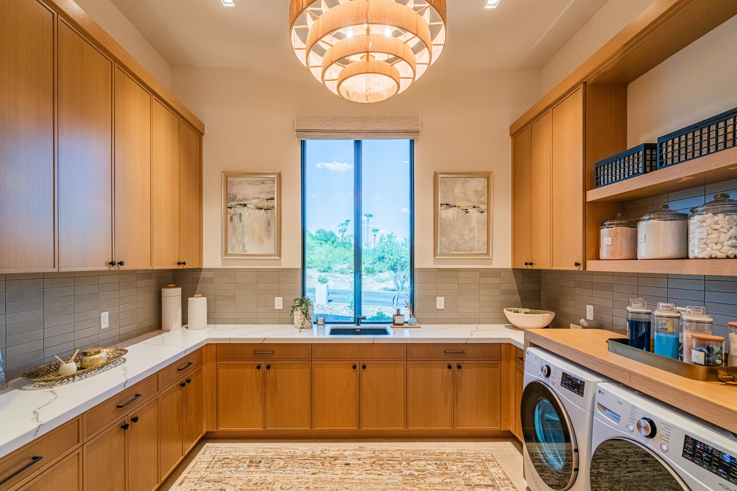 Modern kitchen with wooden cabinets, a window above the sink showing trees outside, a white countertop, and various containers including jars on shelves, with a washer and dryer in the corner.