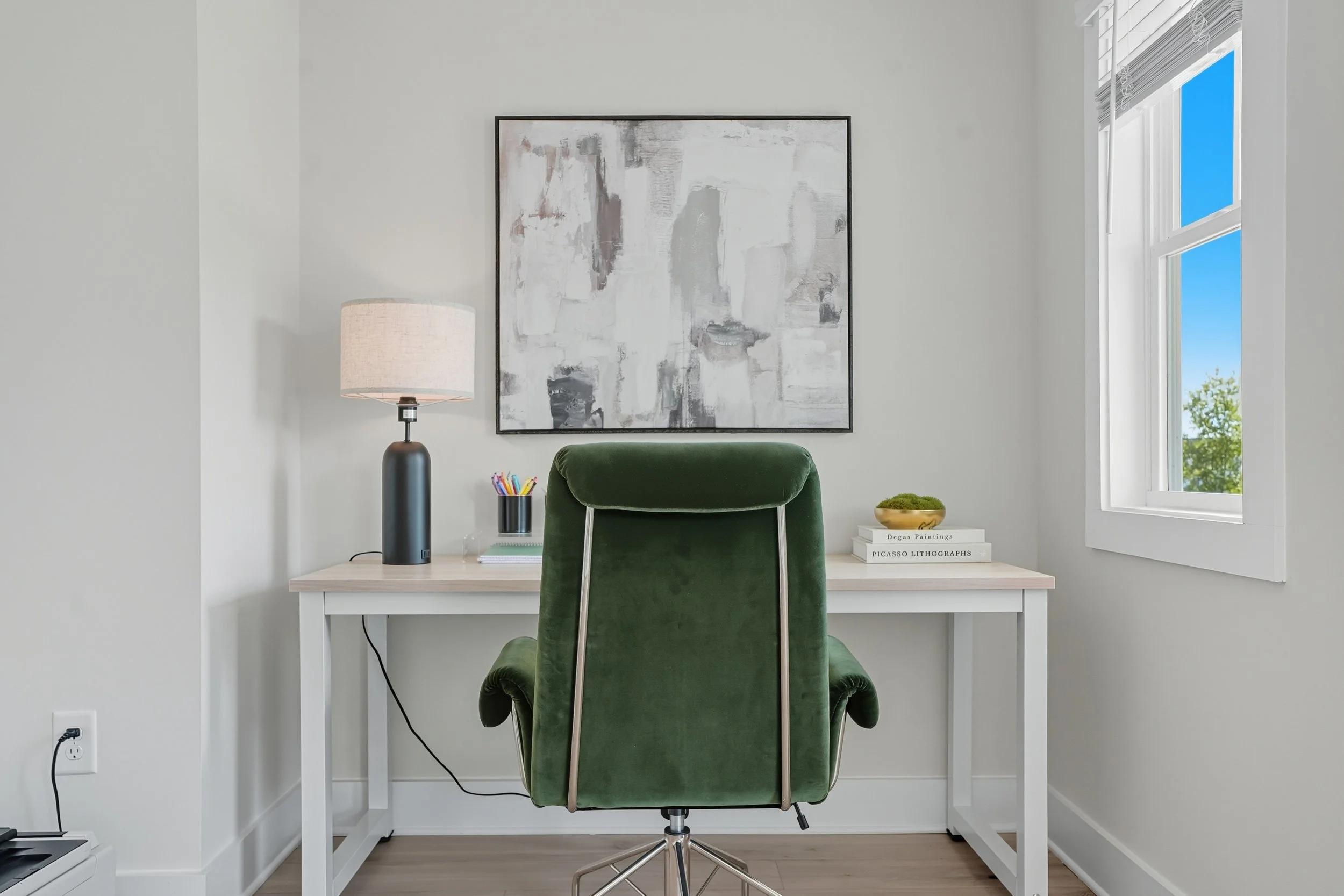 A home office with a white desk, green office chair, abstract black, white, and gray painting on the wall, a black table lamp, books about Picasso lithographs, and a window showing a blue sky.