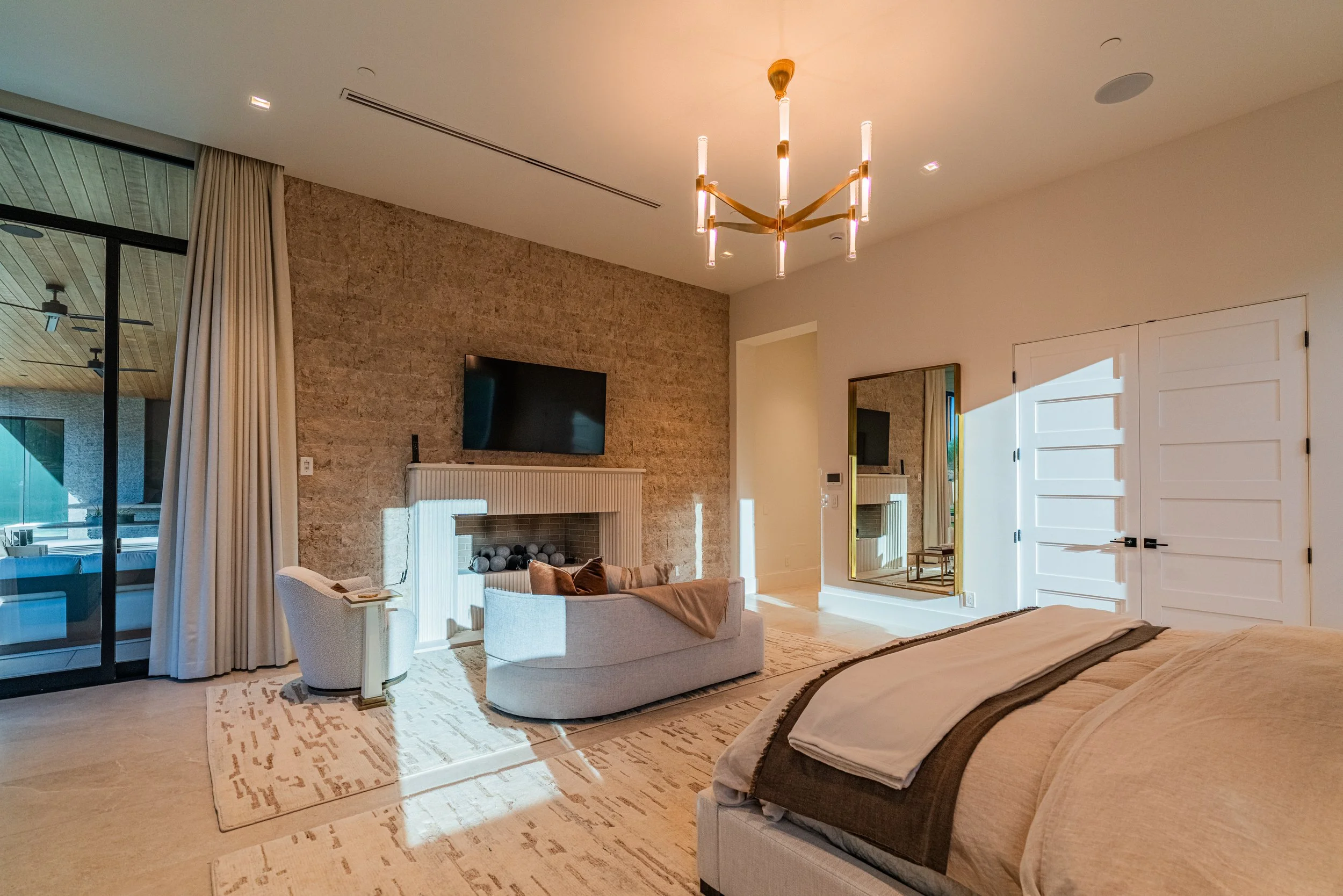 Modern living room with white couch, fireplace, and wall-mounted TV. Large windows with curtains, wooden ceiling, chandelier lighting, and a door with horizontal panels.