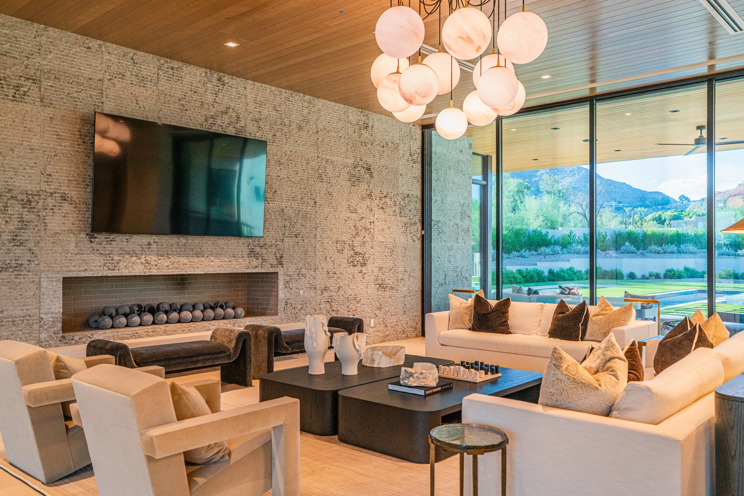 Modern living room with large windows showing outdoor mountains and greenery, a stone fireplace with a flat-screen TV above it, and a chandelier with spherical pendant lights hanging from the ceiling. The room has beige sofas and various decorative c
