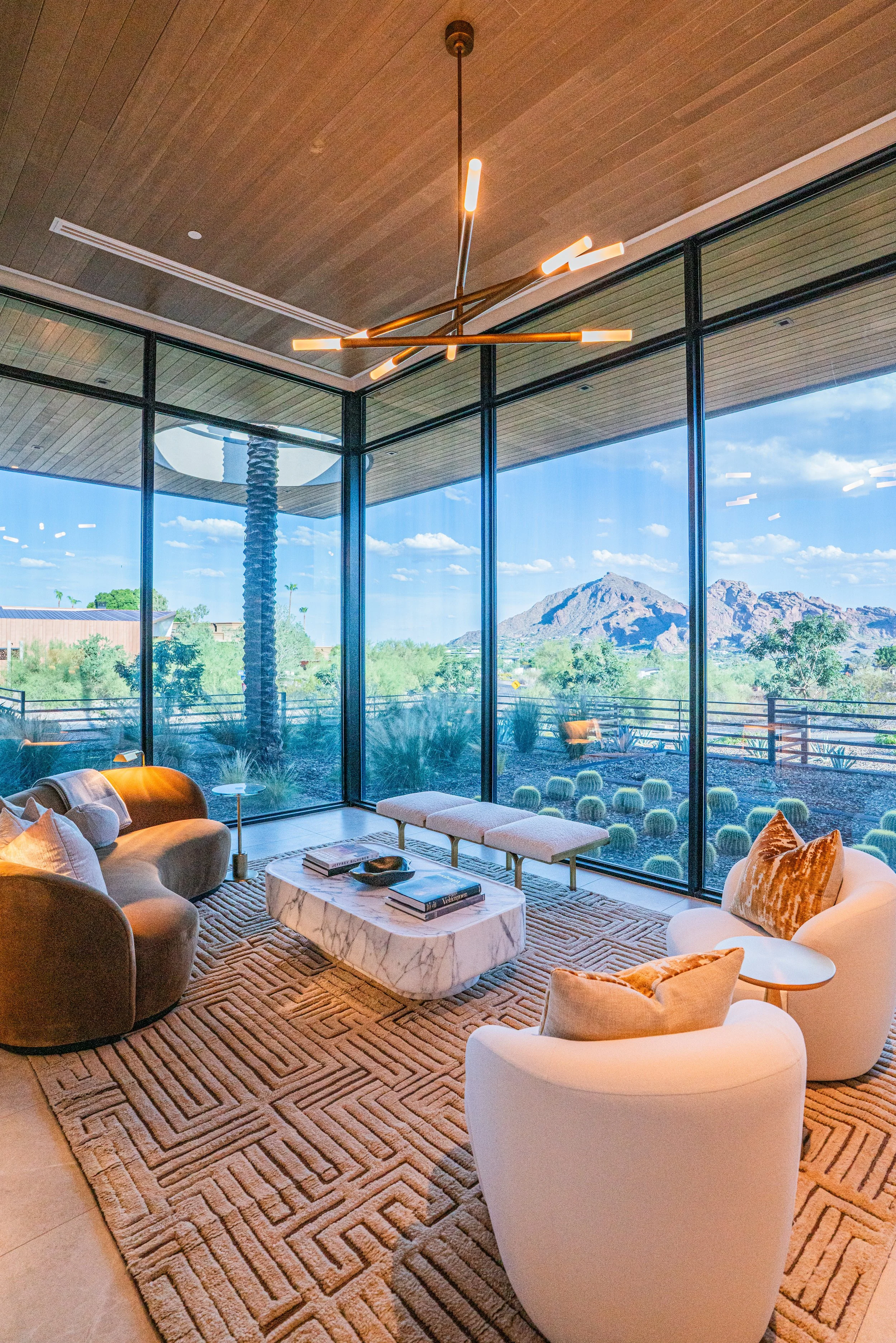Modern living room with floor-to-ceiling glass windows showing mountain and desert landscape, stylish beige and brown furniture, marble coffee table, warm lighting, and a patterned carpet.