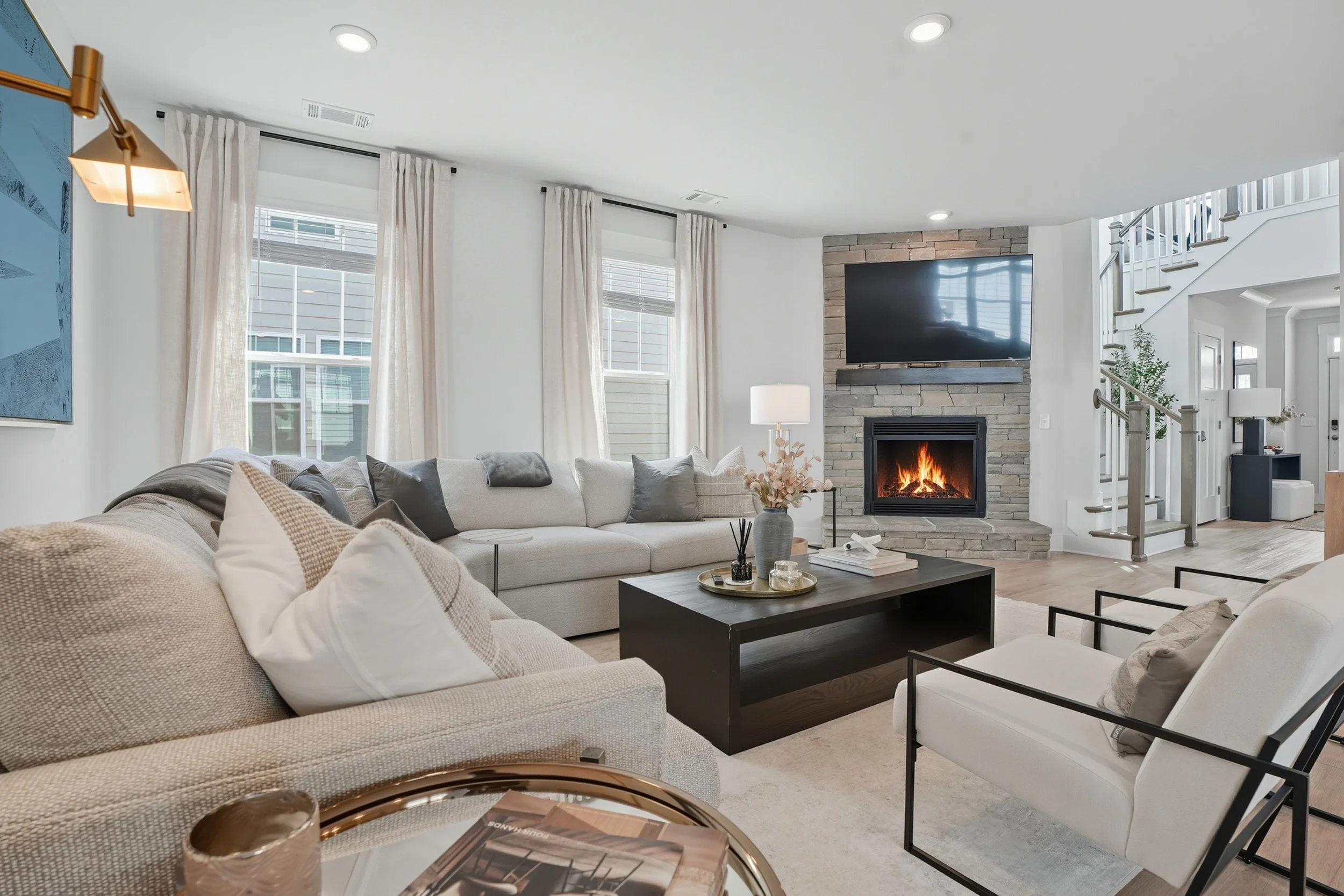 Modern living room with beige sectional sofa, coffee table, fireplace with mounted TV, large windows with white curtains, and a staircase in the background.