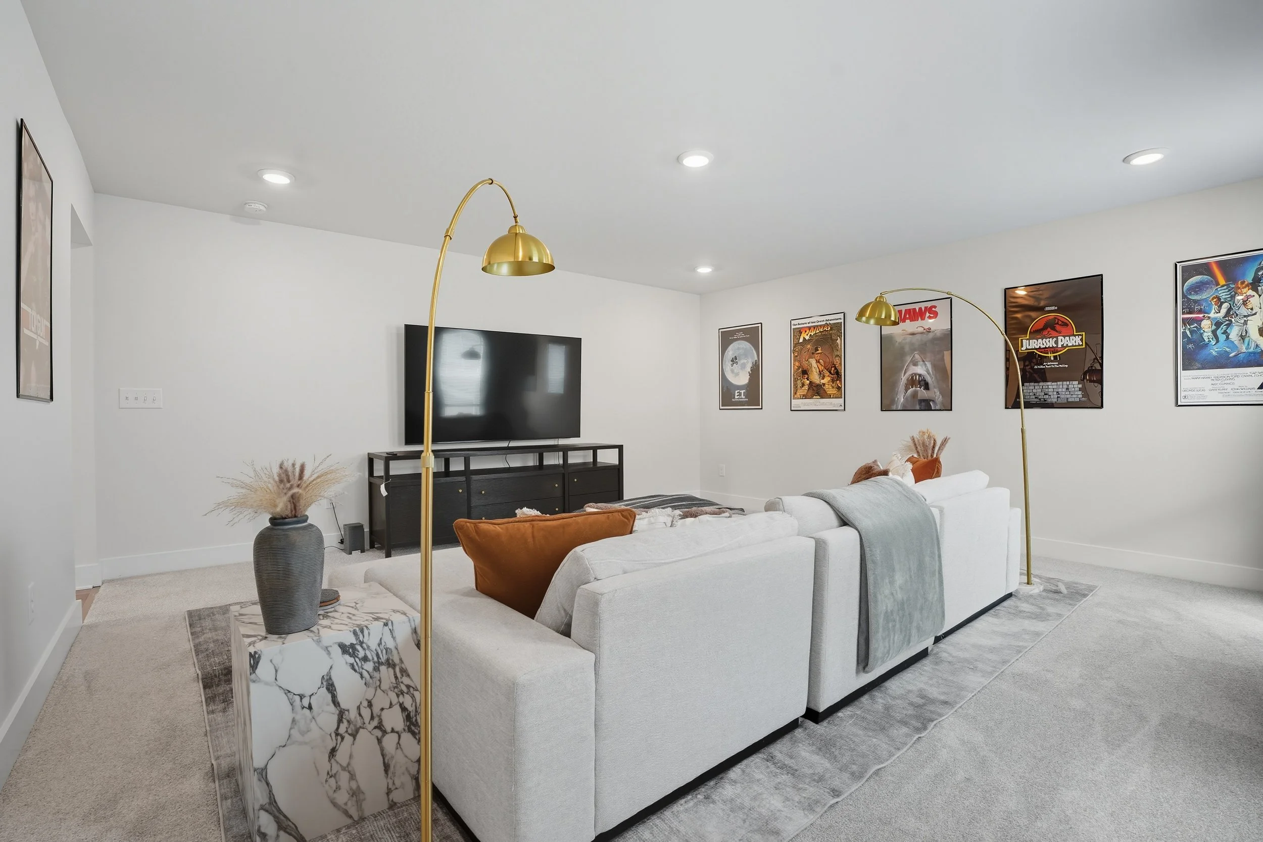 Modern living room with a white sofa, gold floor lamps, a black entertainment center with a television, and framed movie posters on white walls.