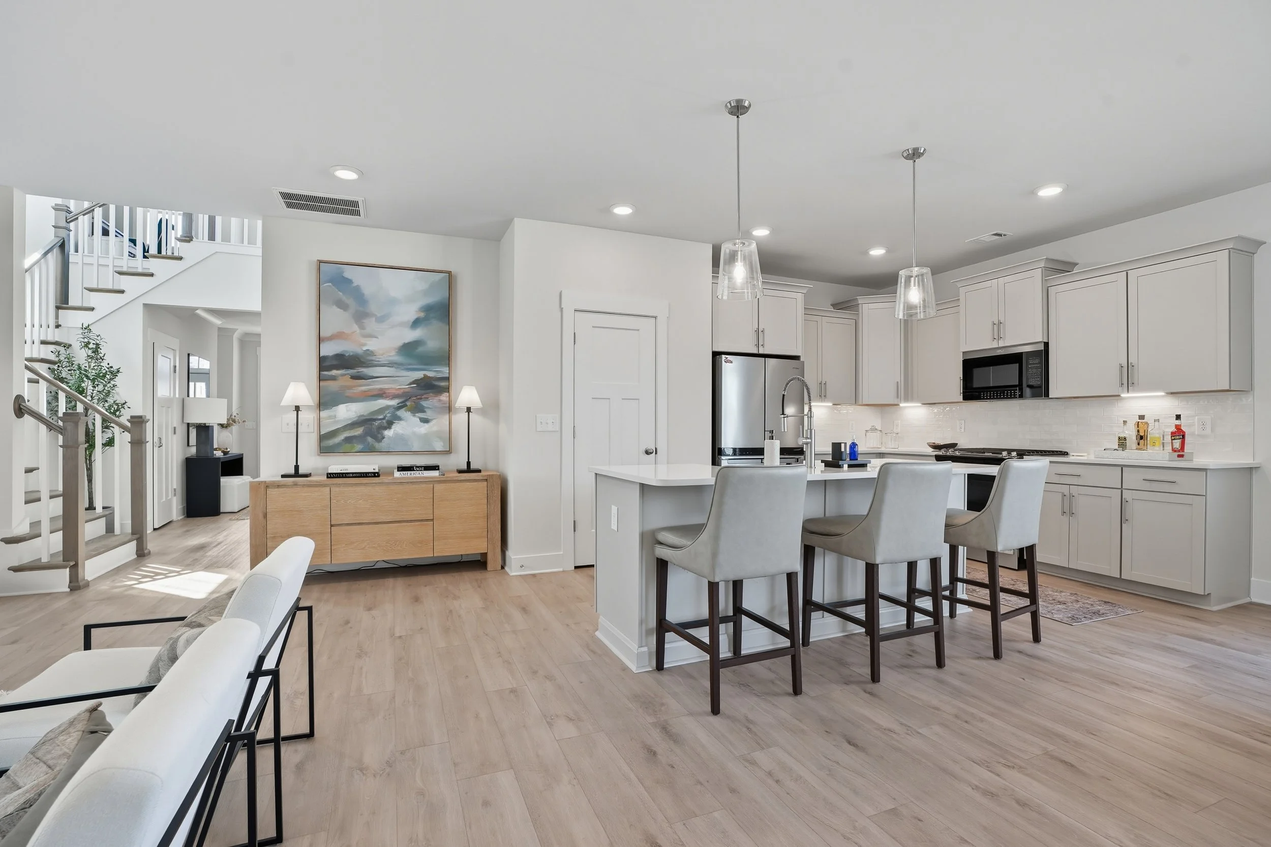 Open-concept kitchen and living area with white cabinetry, a kitchen island with four beige chairs, a stainless steel refrigerator, black microwave, painting, and staircase in the background.