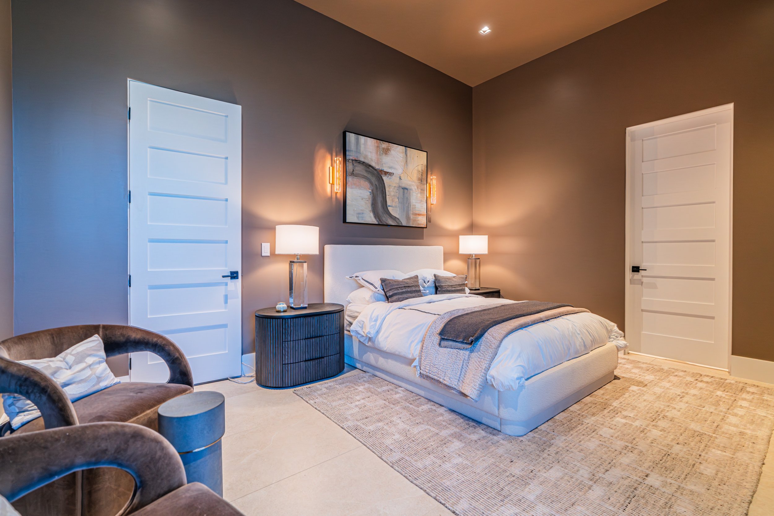 Modern bedroom with a white bed, gray walls, two white doors, abstract artwork above the bed, and two bedside lamps illuminating the room.