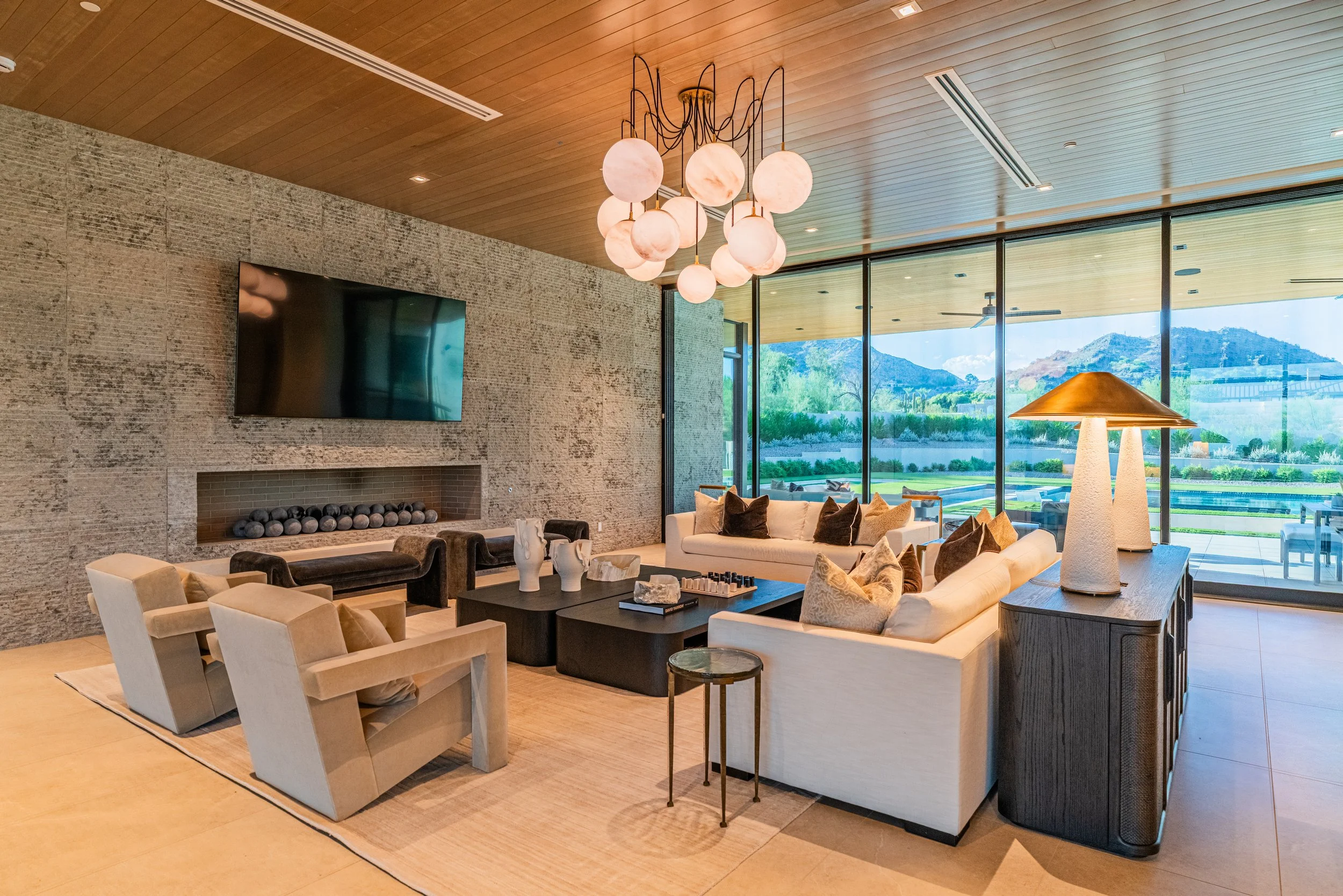 Modern living room with beige sofas, a black coffee table, a textured brick fireplace, a large flat-screen TV, and floor-to-ceiling windows showing a scenic outdoor landscape with mountains and greenery. Elegant light fixtures hang from a wooden ceil