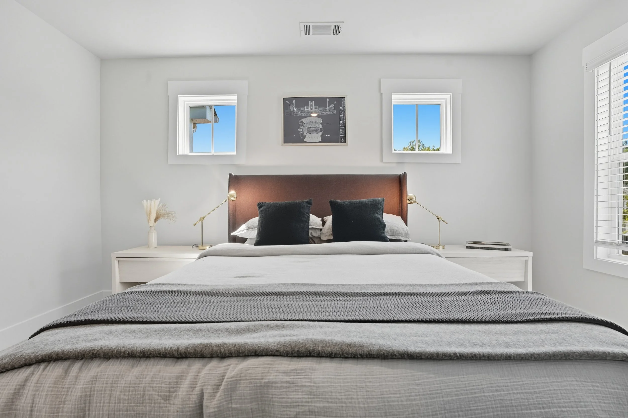 A neatly made bed with a gray blanket and black pillows in a minimalist white bedroom with two windows and a framed blueprint on the wall.