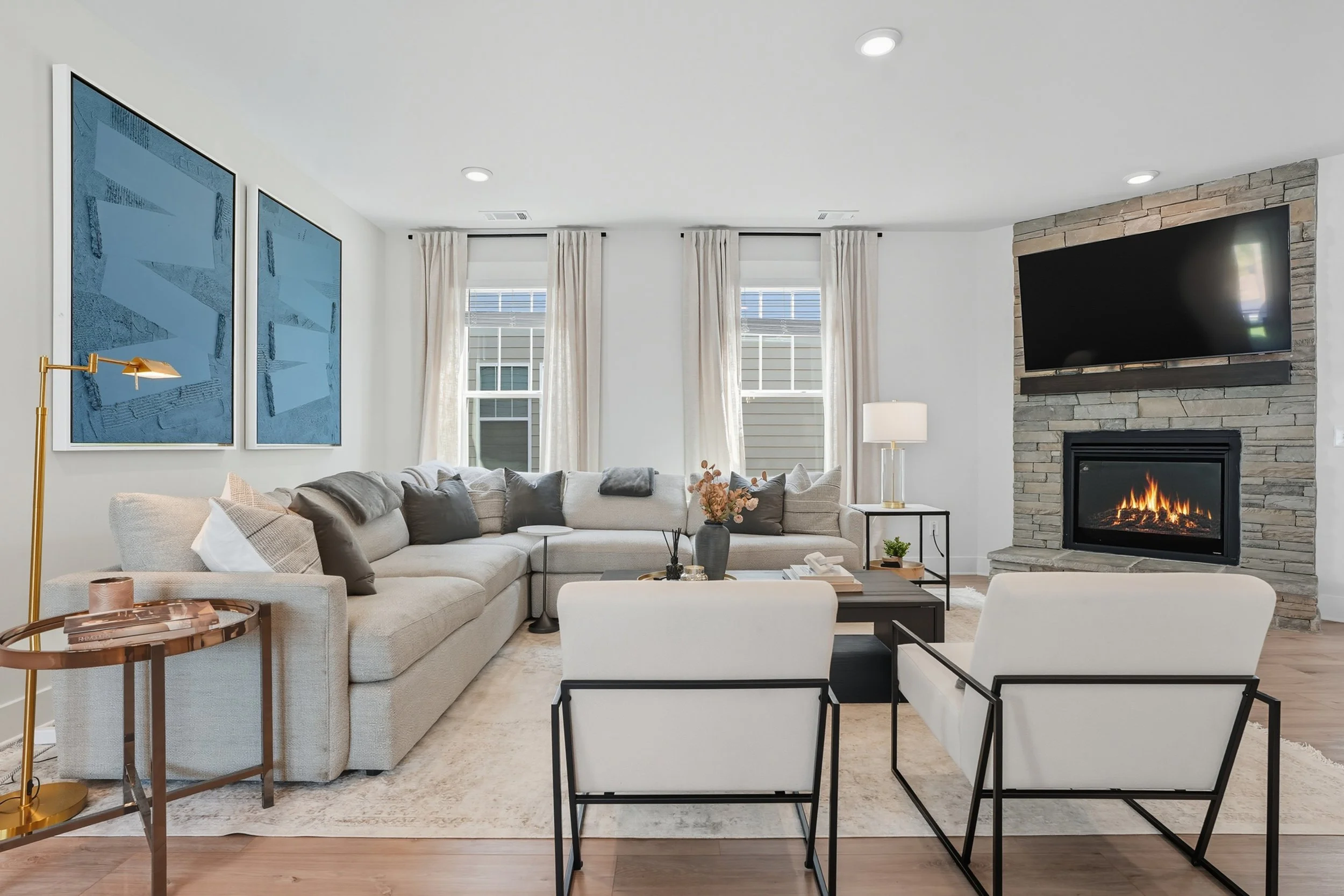 A modern living room with a sectional sofa, armchair, fireplace, wall-mounted TV, artwork, and large windows with white curtains.