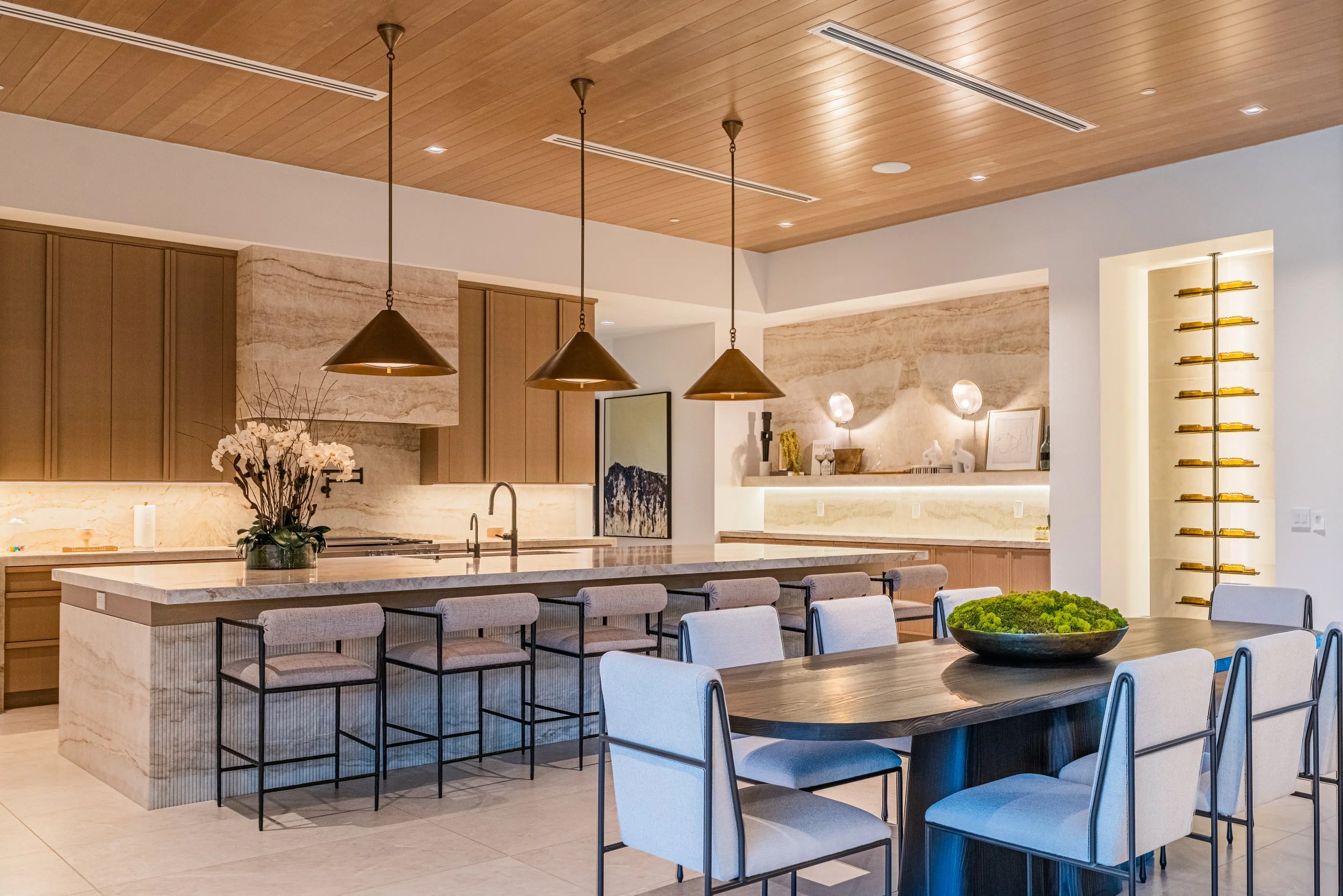 Modern kitchen and dining area with wooden ceiling, pendant lights, and a large marble island with chairs, white walls, and decorative shelves.