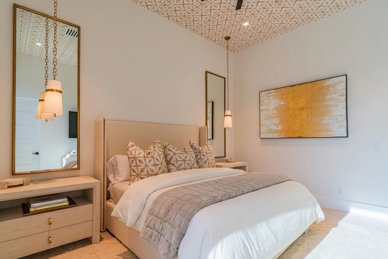 Modern bedroom with beige upholstered bed, white bedding, beige and geometric patterned pillows, light wood nightstands, large mirror with gold frame, abstract yellow and white painting, pendant lights with white shades, and patterned ceiling design.