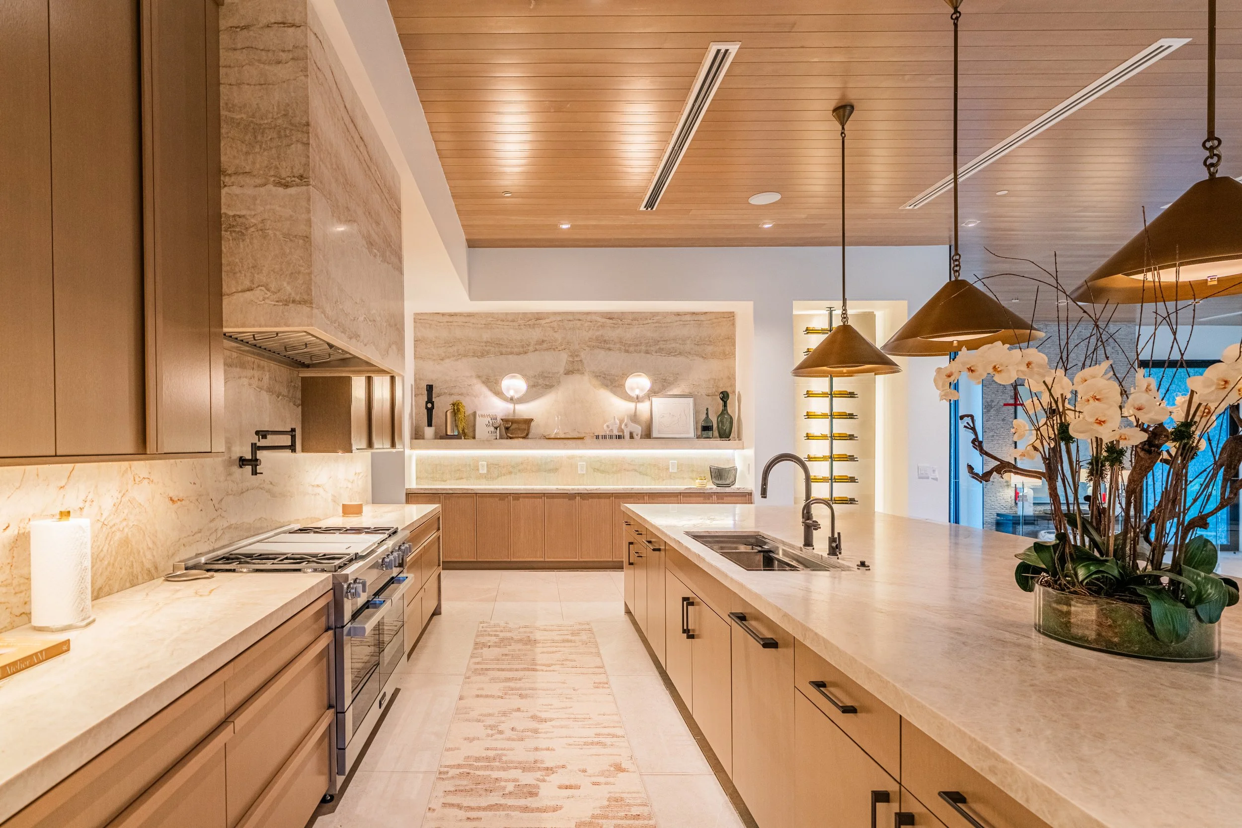 A modern kitchen with light wood cabinets, a marble countertop, pendant lights, and decorative items on a shelf. A large island with a built-in sink is in the foreground, and there are flowers and plants on the right side.