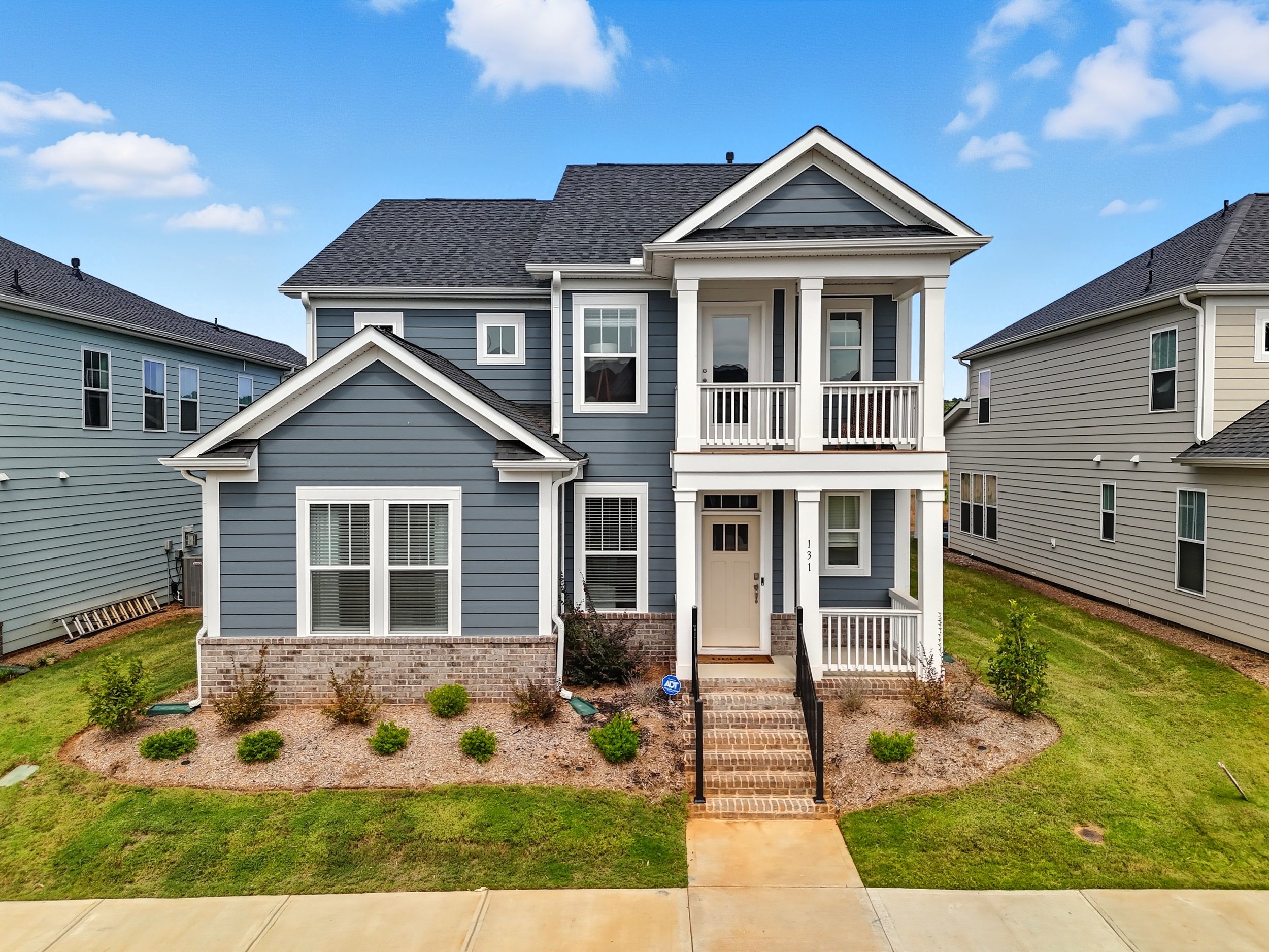 Front view of a two-story blue house with white trim, brick stairs leading to the front door, and a small landscaped yard with bushes and grass, surrounded by neighboring houses, under a blue sky with scattered clouds.