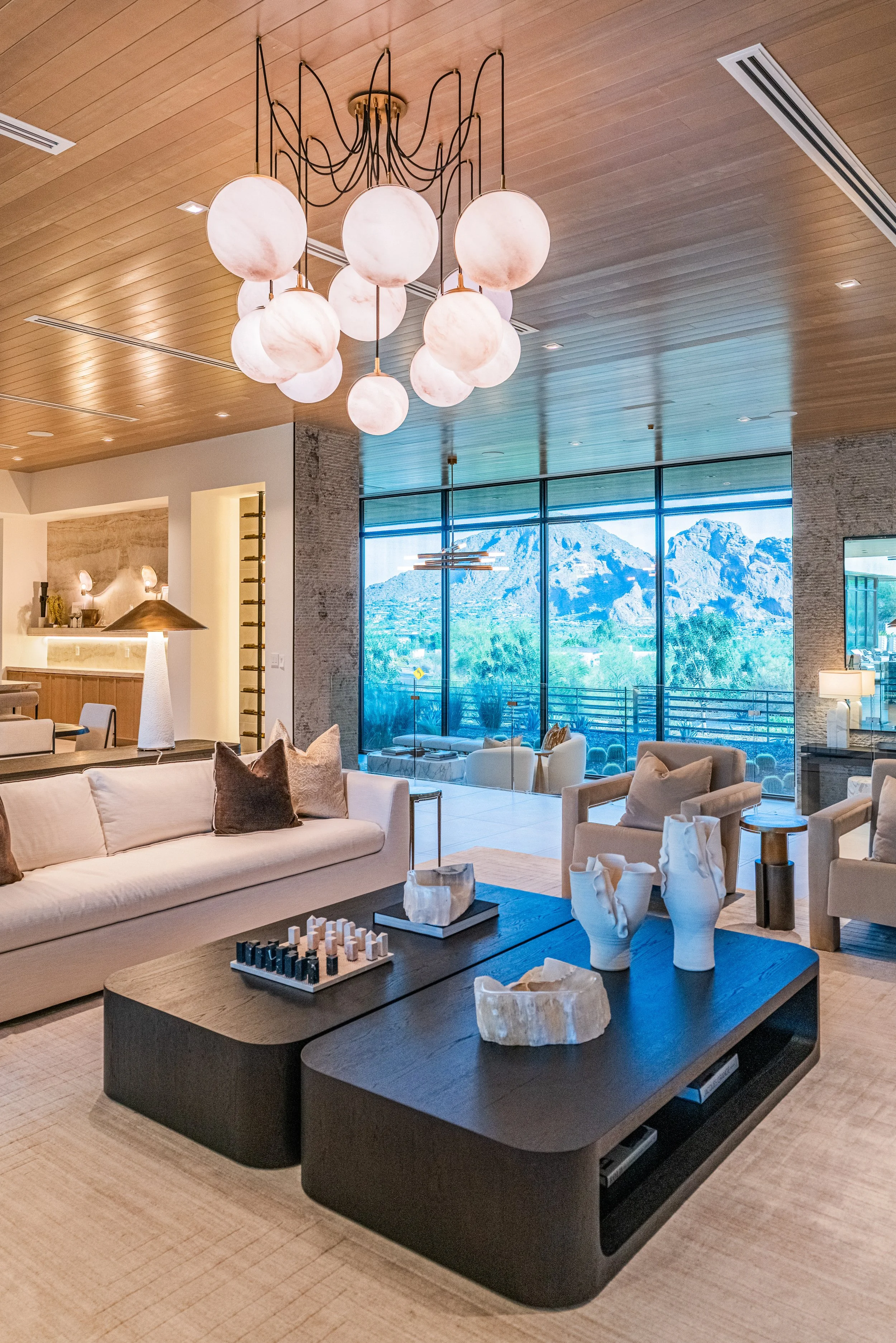 Modern living room with beige sofa, dark coffee table with chess set and sculptures, white armchair, large window showing mountains, and wooden ceiling with pendant lighting.