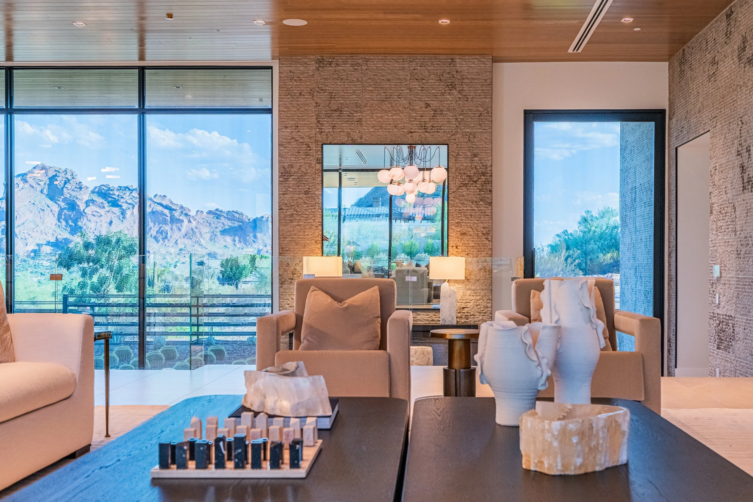 Interior of a modern living room with large windows showcasing mountains and blue sky, beige and white furniture, a chandelier, and a chessboard on the black coffee table.