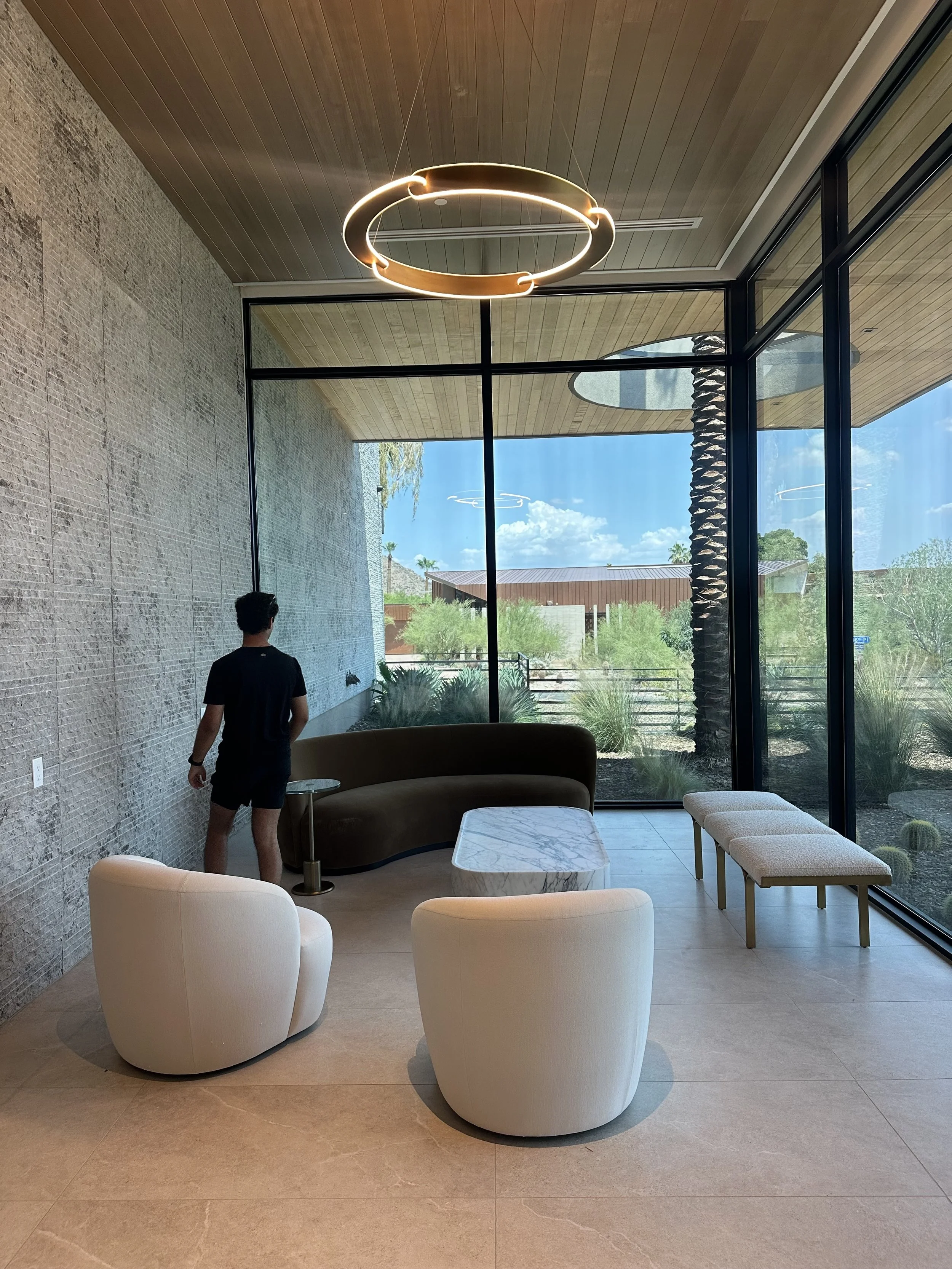Modern lounge area with white curved chairs, marble table, large floor-to-ceiling windows, and a view of a patio with trees and a clear blue sky.