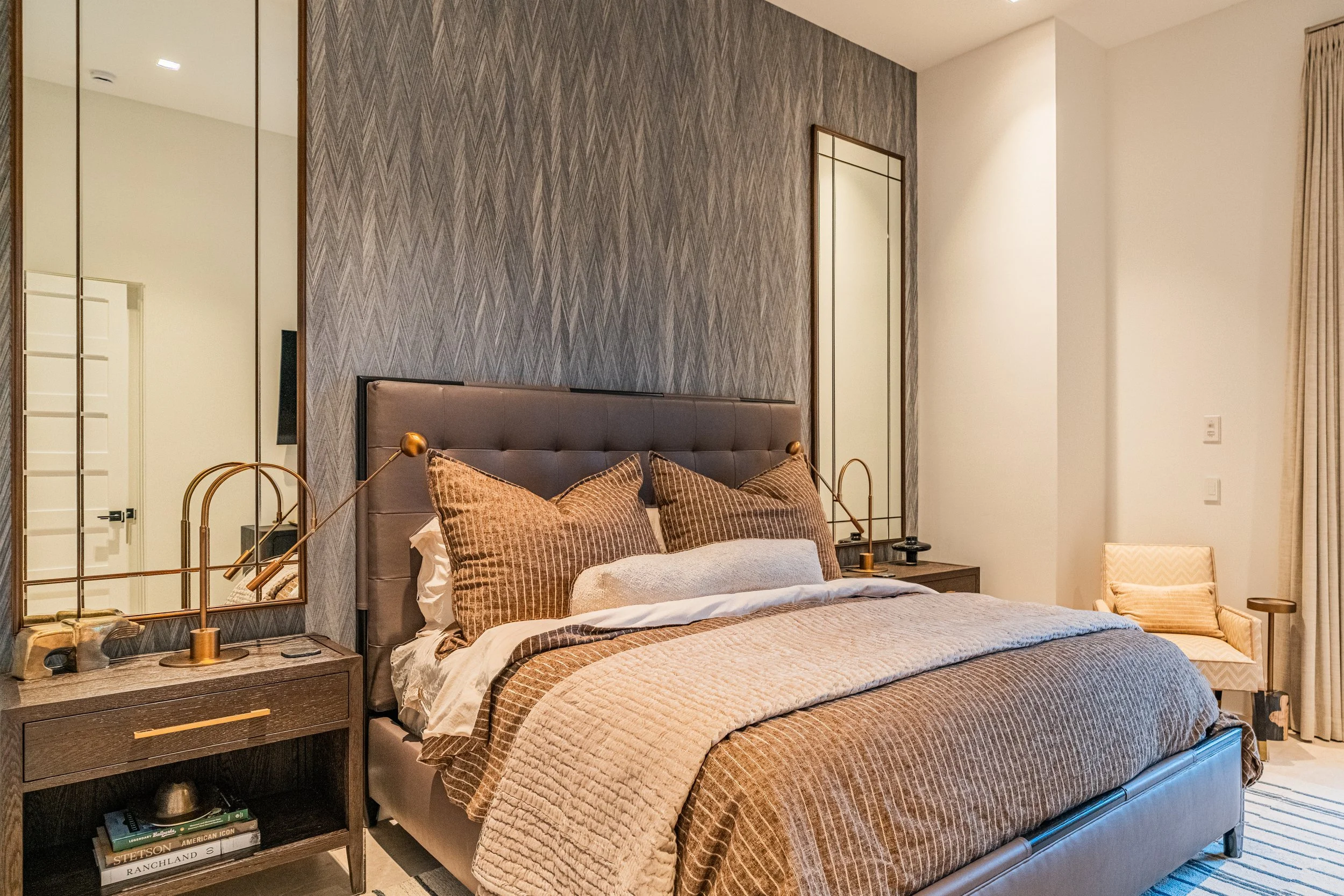 A cozy bedroom with a large bed featuring brown and beige bedding and pillows. There are two wooden nightstands with modern lamps, a decorative chair in the corner, a patterned wall behind the bed, and a large mirror on the wall.