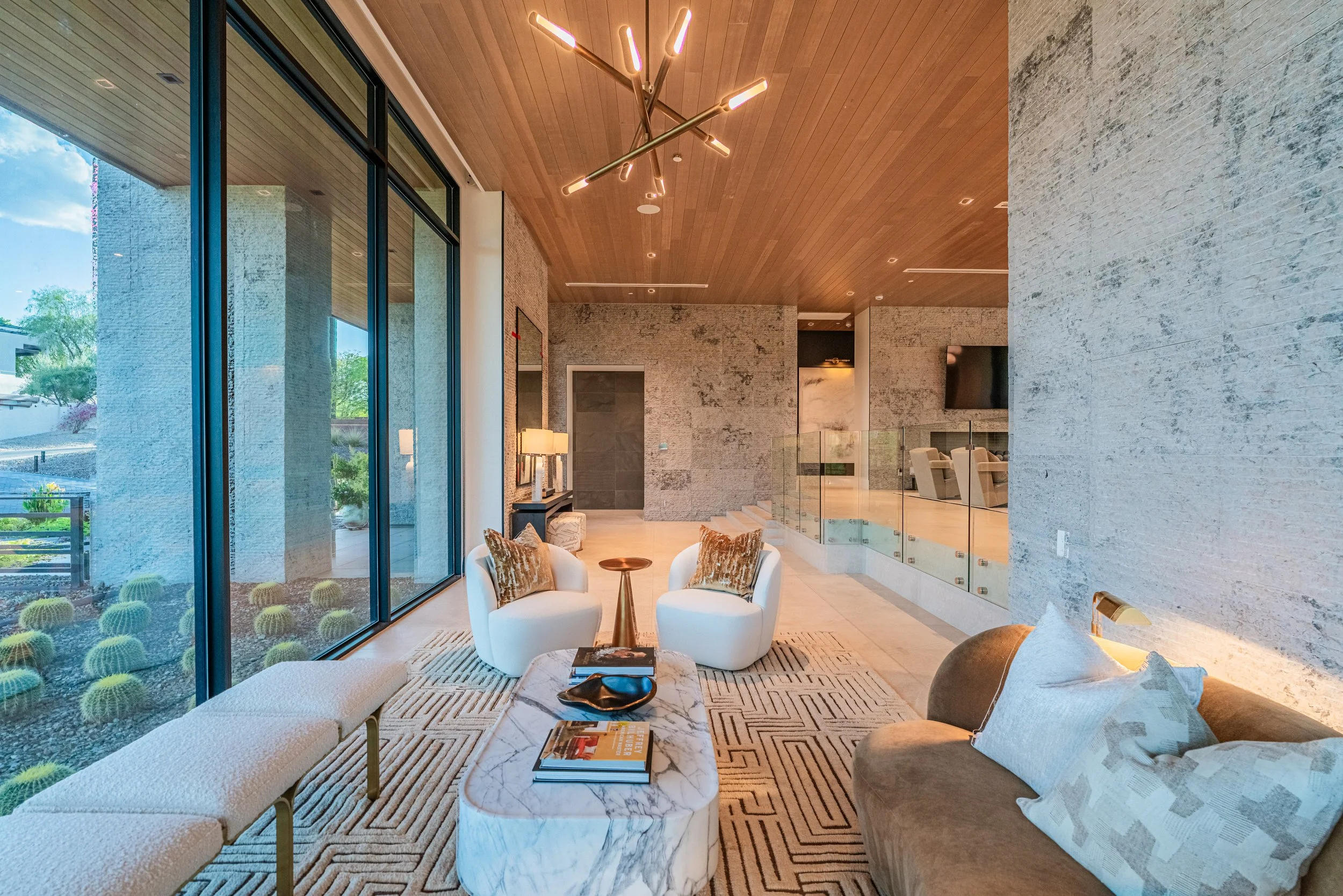Modern living room with large floor-to-ceiling glass windows, a wood-paneled ceiling, and contemporary furniture including white armchairs, a marble coffee table, and a sofa with pillows. There are books and decorative items on the coffee table, and 