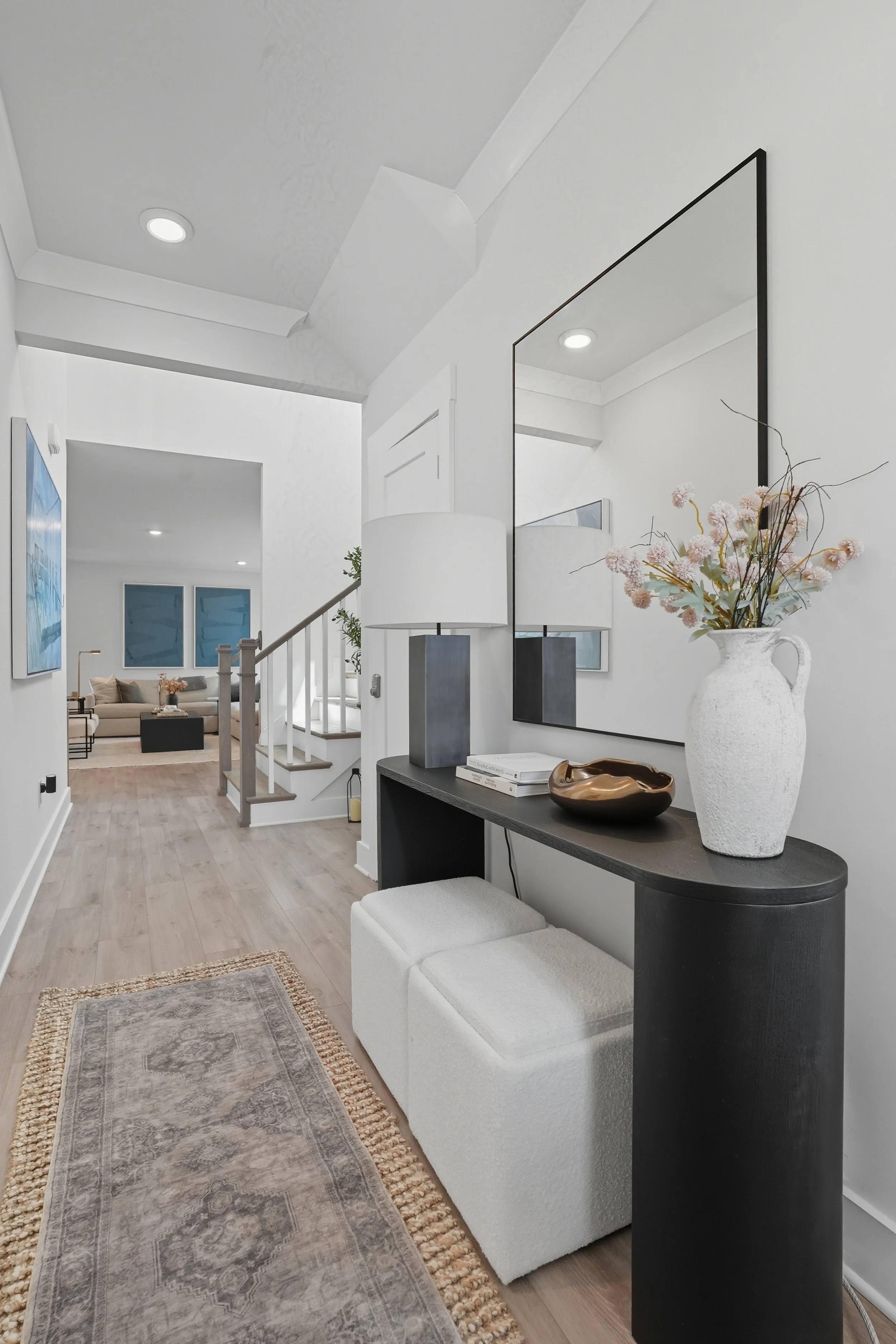 Interior view of a modern living space featuring a console table with a large white ceramic vase holding pink flowers, a decorative bowl, and a table lamp. The room has light wood flooring, a white couch, artwork on the walls, and a staircase leading
