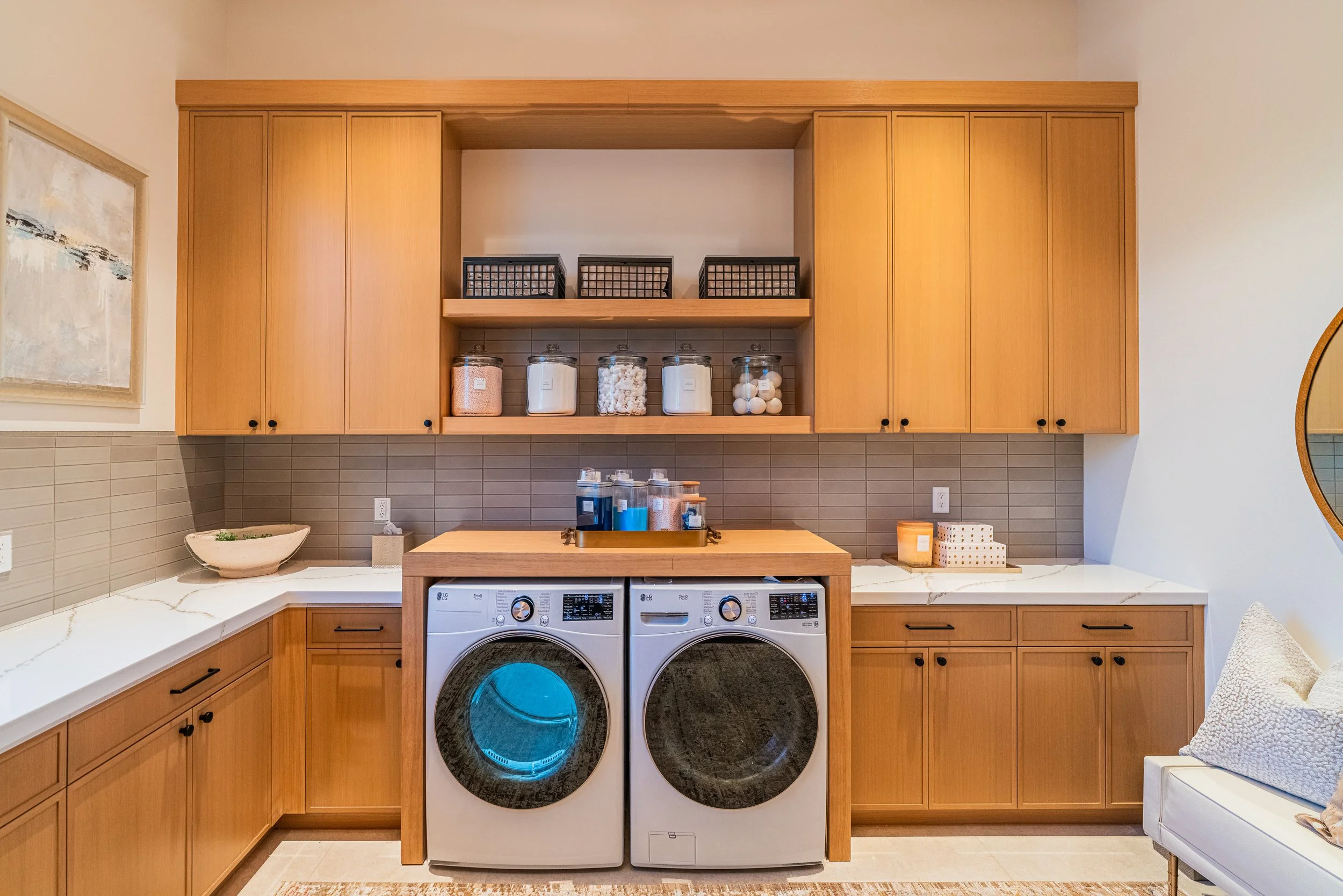 Laundry room with wooden cabinets, a countertop with a bowl, and a washing machine and dryer with digital controls, laundry baskets on shelves, and a mirror on the right side.