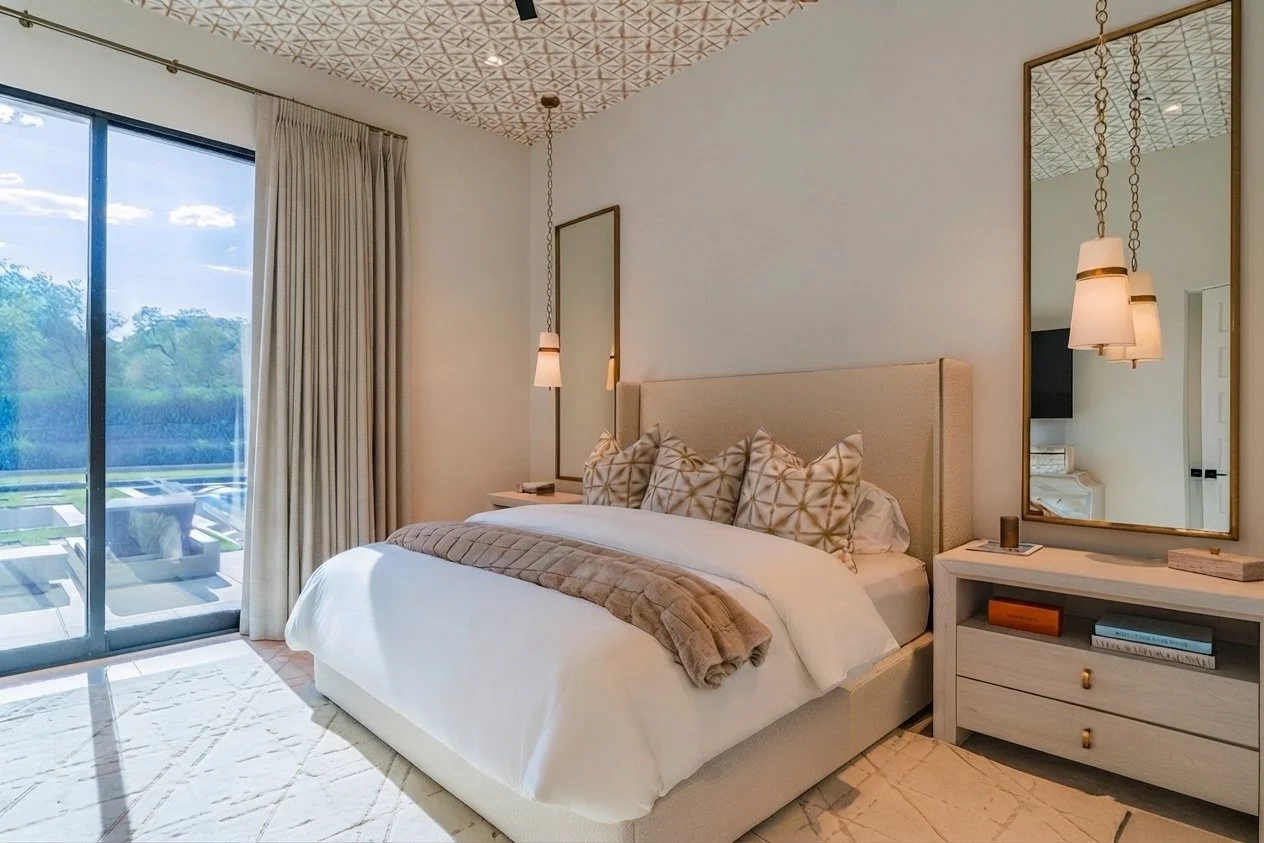 Elegant bedroom with a large bed, multiple patterned pillows, a beige headboard, and a white duvet. A cozy blanket is draped over the foot of the bed. Two mirrors hang on the white walls, and a modern bedside table with books and a decorative item is