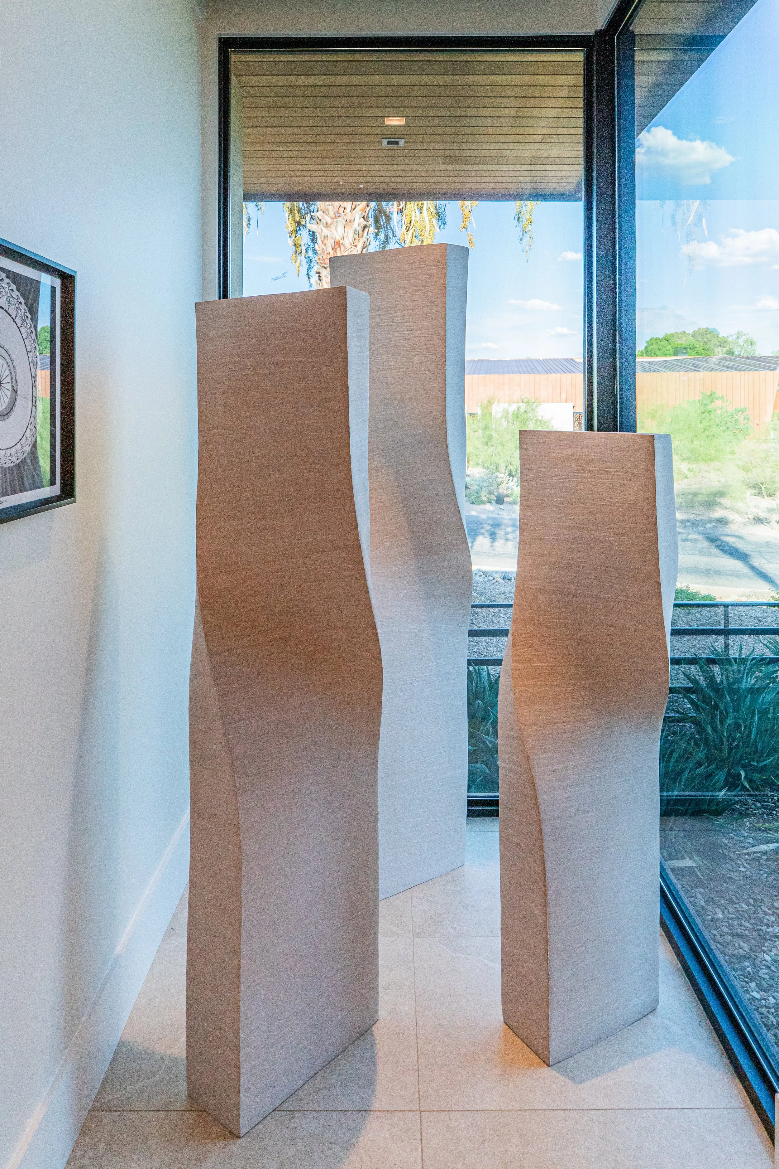 Three tall, light-colored wooden sculptures with curved designs are positioned near a large window showing trees, sky, and a building outside. There is a framed picture on the left wall.