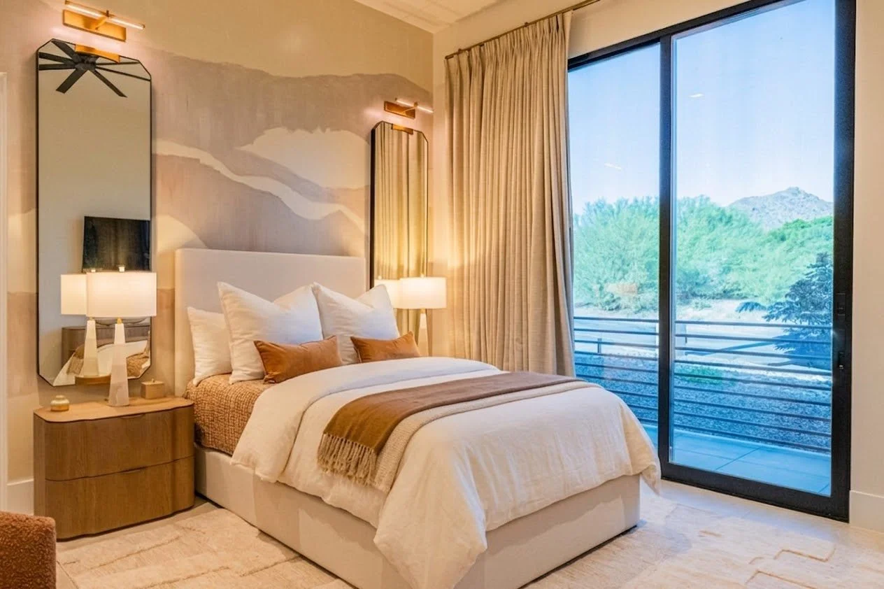 A modern bedroom with a bed, twin bedside tables, lamps, and large sliding glass doors overlooking a balcony with a scenic outdoor view of trees and mountains.