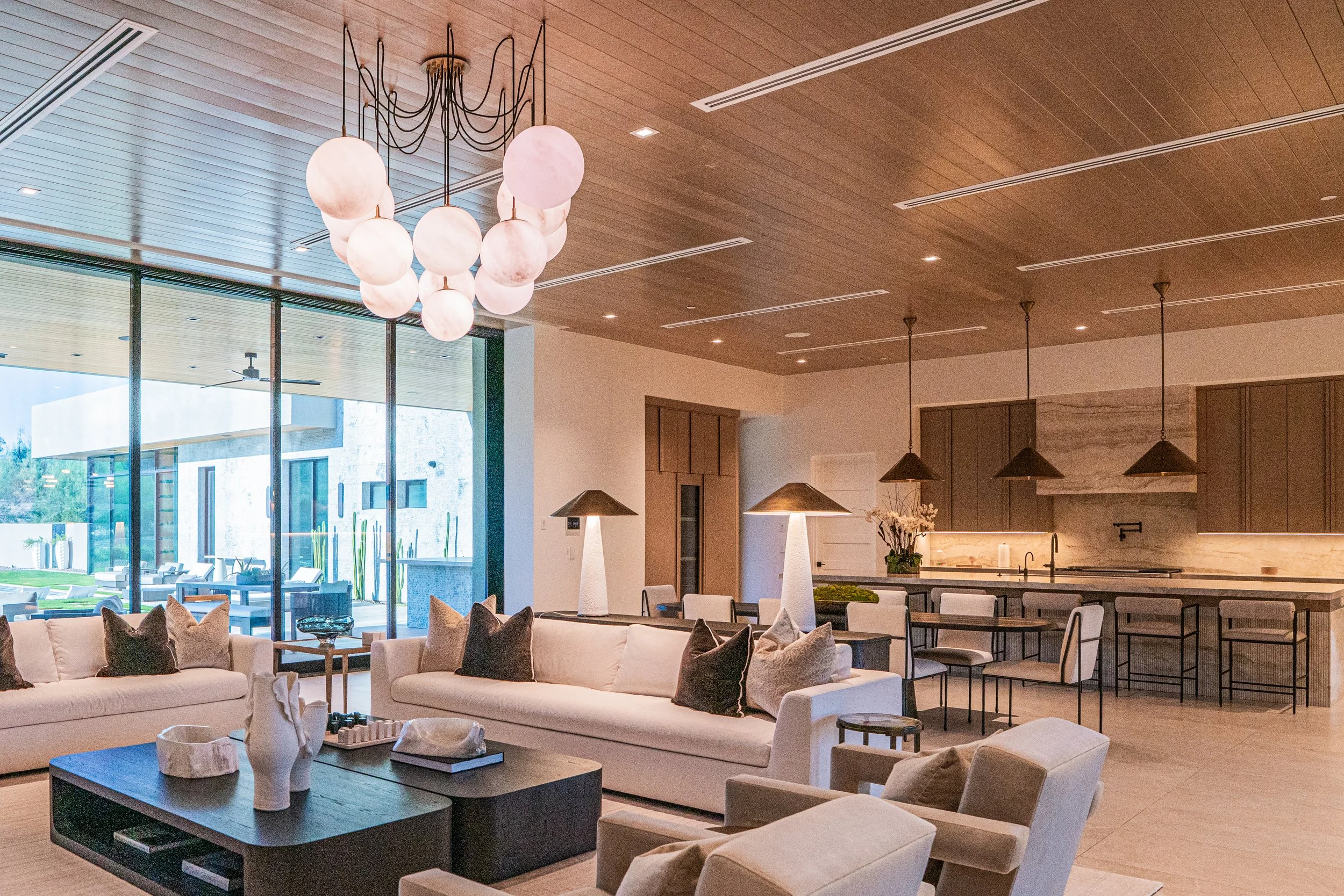 Modern living room with large window, white sofa with brown and black pillows, central dark coffee table with decorative items, abstract lighting fixture with multiple round glass orbs, and a kitchen island in the background with pendant lights.