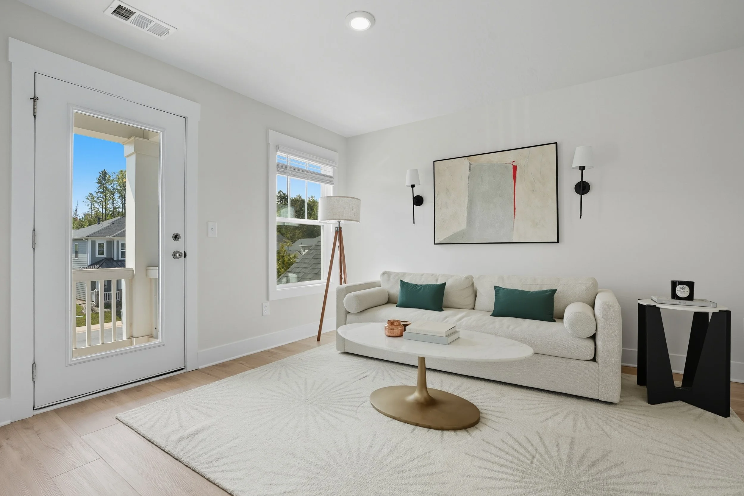 Bright living room with white walls, a beige sofa with green pillows, a white coffee table, a framed abstract art piece, a tall floor lamp, wall sconces, and a door leading outside showing a view of neighboring houses and trees.