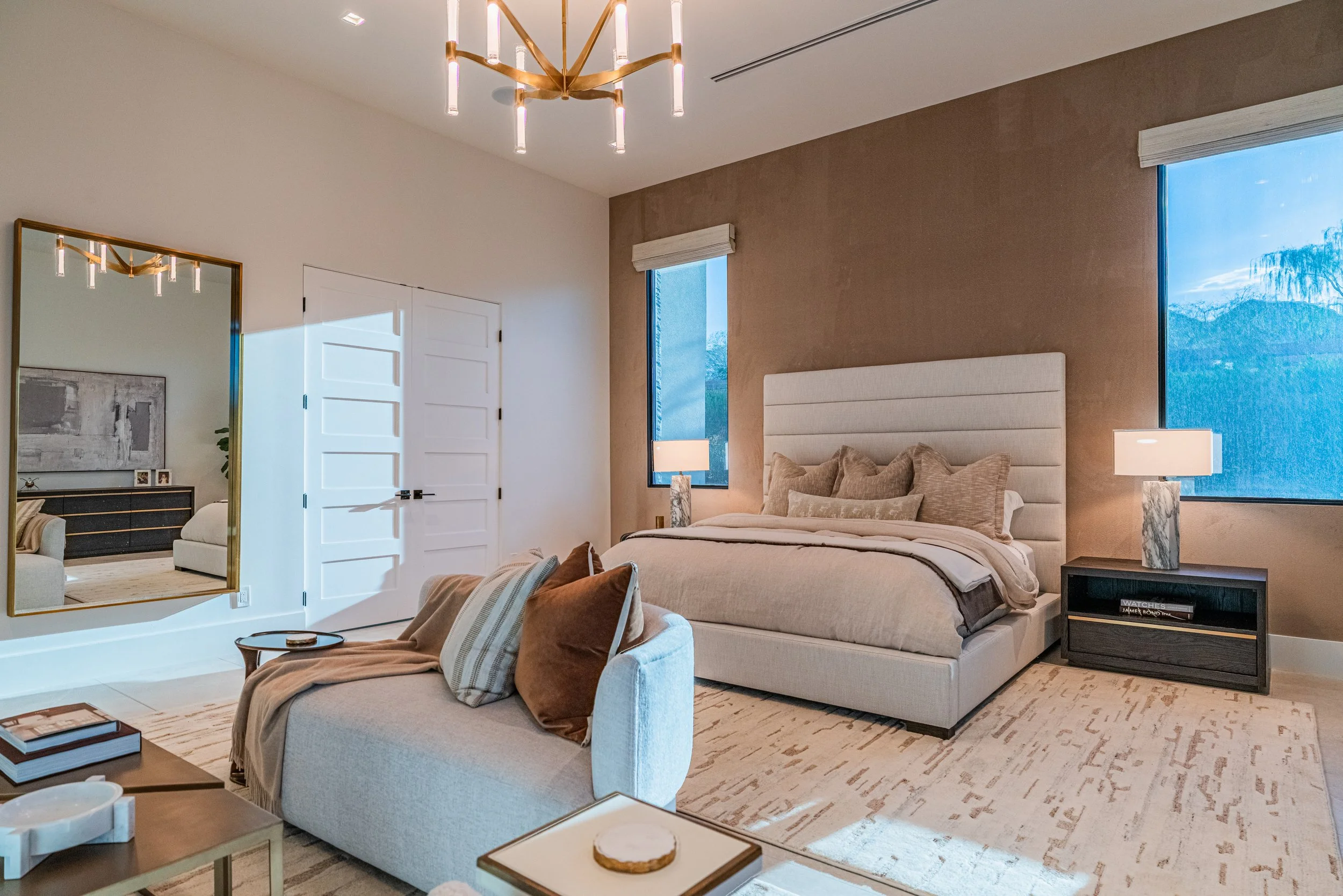 A modern bedroom with a large bed, beige headboard, and beige bedding; two nightstands with lamps, large windows showing a view of trees, a white closet door, and a ceiling chandelier with multiple light fixtures.
