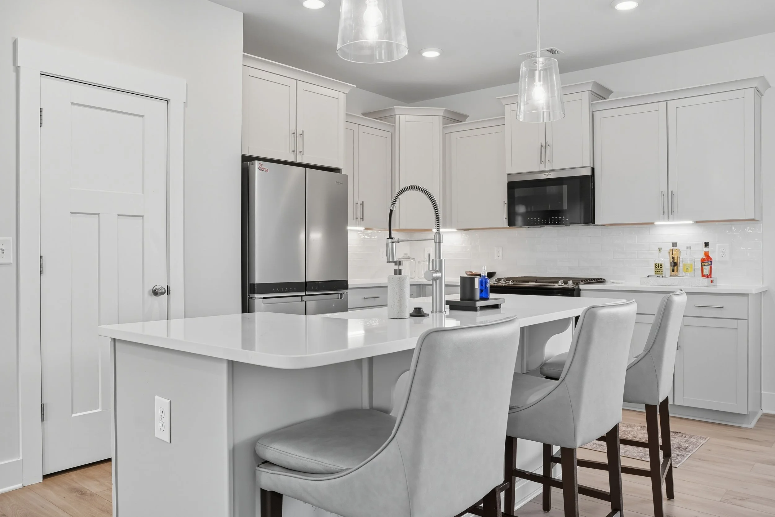 Modern white kitchen with island, stainless steel refrigerator, built-in microwave, white cabinets, and three pendant lights, with a dining table and chairs in the foreground.