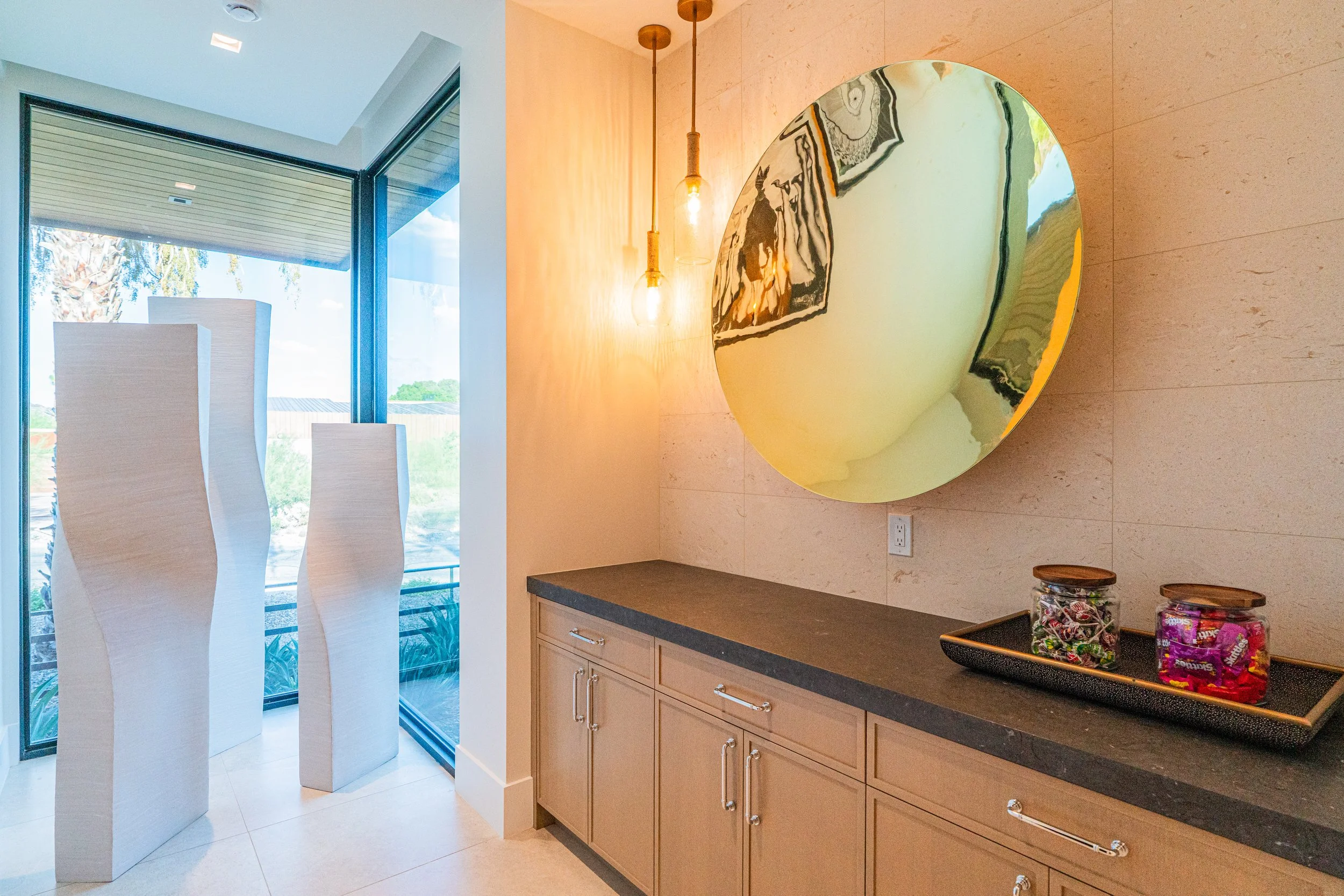 Interior view of a modern kitchen with beige cabinets, a dark countertop, and decorative jars of candies. A large circular mirror with a marbled pattern is mounted on the wall, and two pendant lights hang from the ceiling. Large windows let in natura