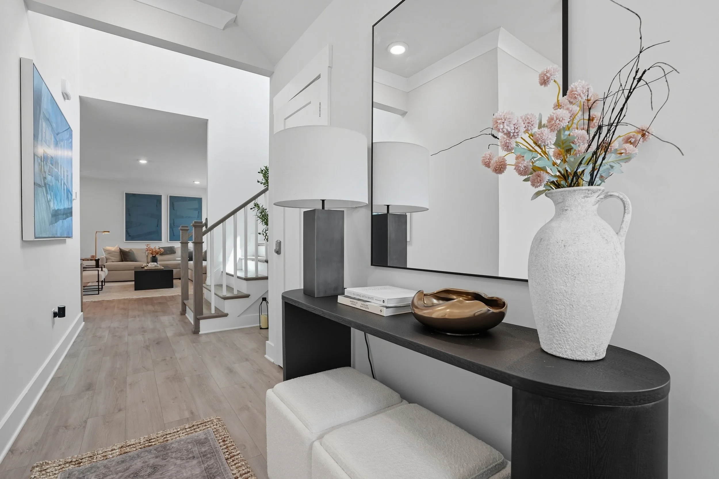 Modern living room with white walls, wooden flooring, and abstract wall art. A front console table holds a large white textured vase with pink flowers and black twigs, a metallic bowl, and books. In the background, comfortable seating with throw pillows and large windows creating natural light.