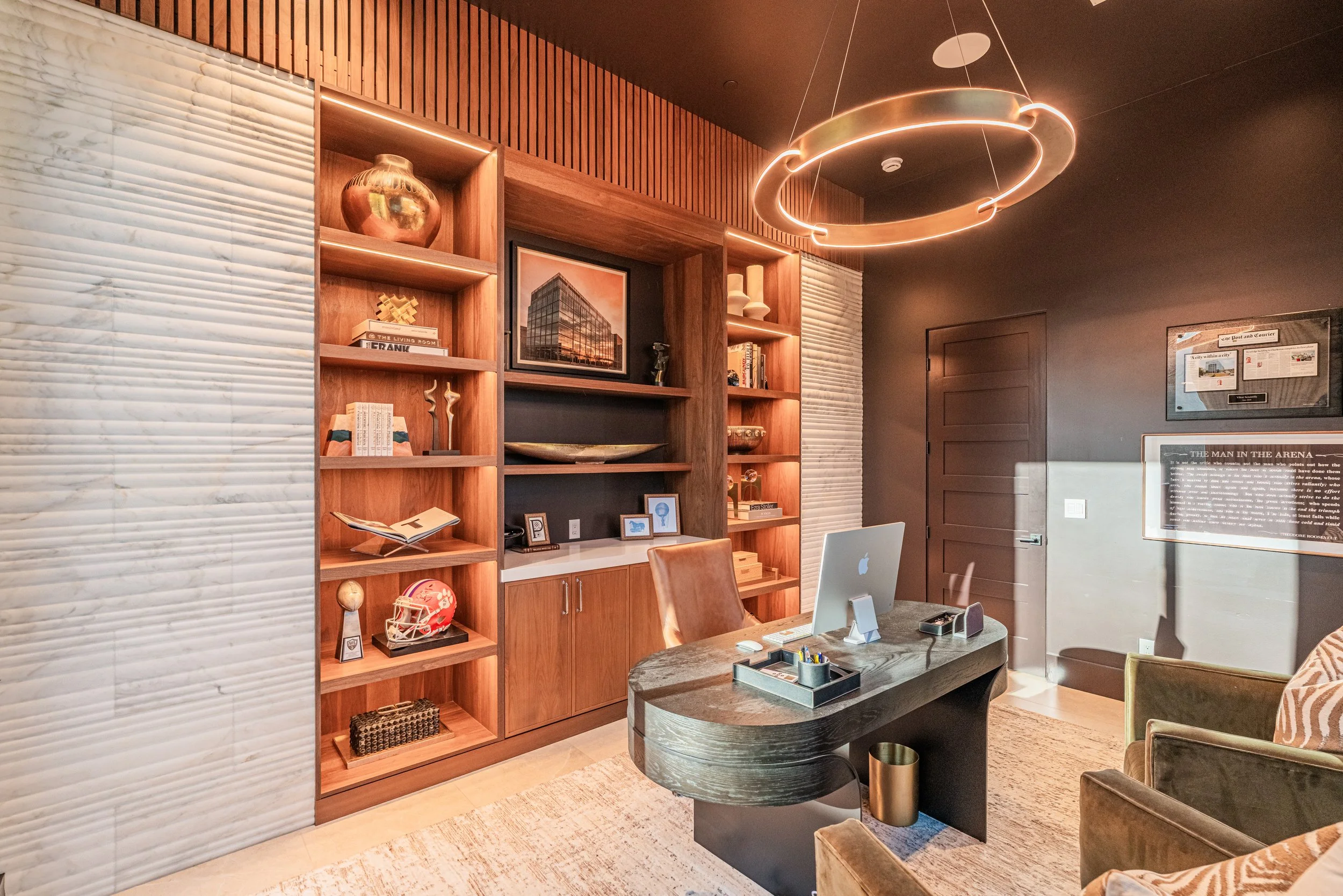 A modern office space with a large wooden bookcase filled with decor, books, and framed photos. There is a desk with an Apple iMac, a leather chair, and a circular black coffee table. A decorative circular light fixture hangs from the ceiling, and th